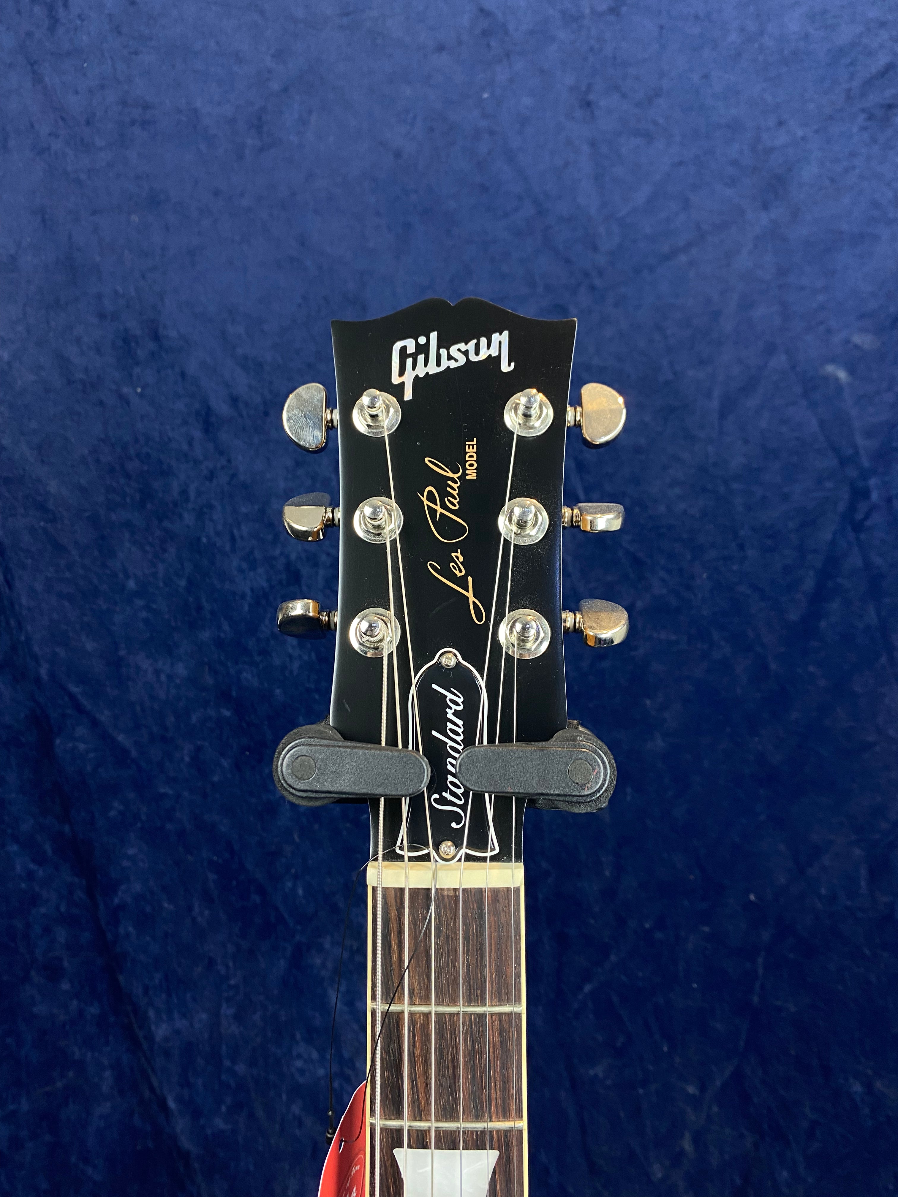Headstock of a Gibson Les Paul Standard electric guitar with gold and white lettering, six tuning pegs, and a dark wood fretboard against a blue background.