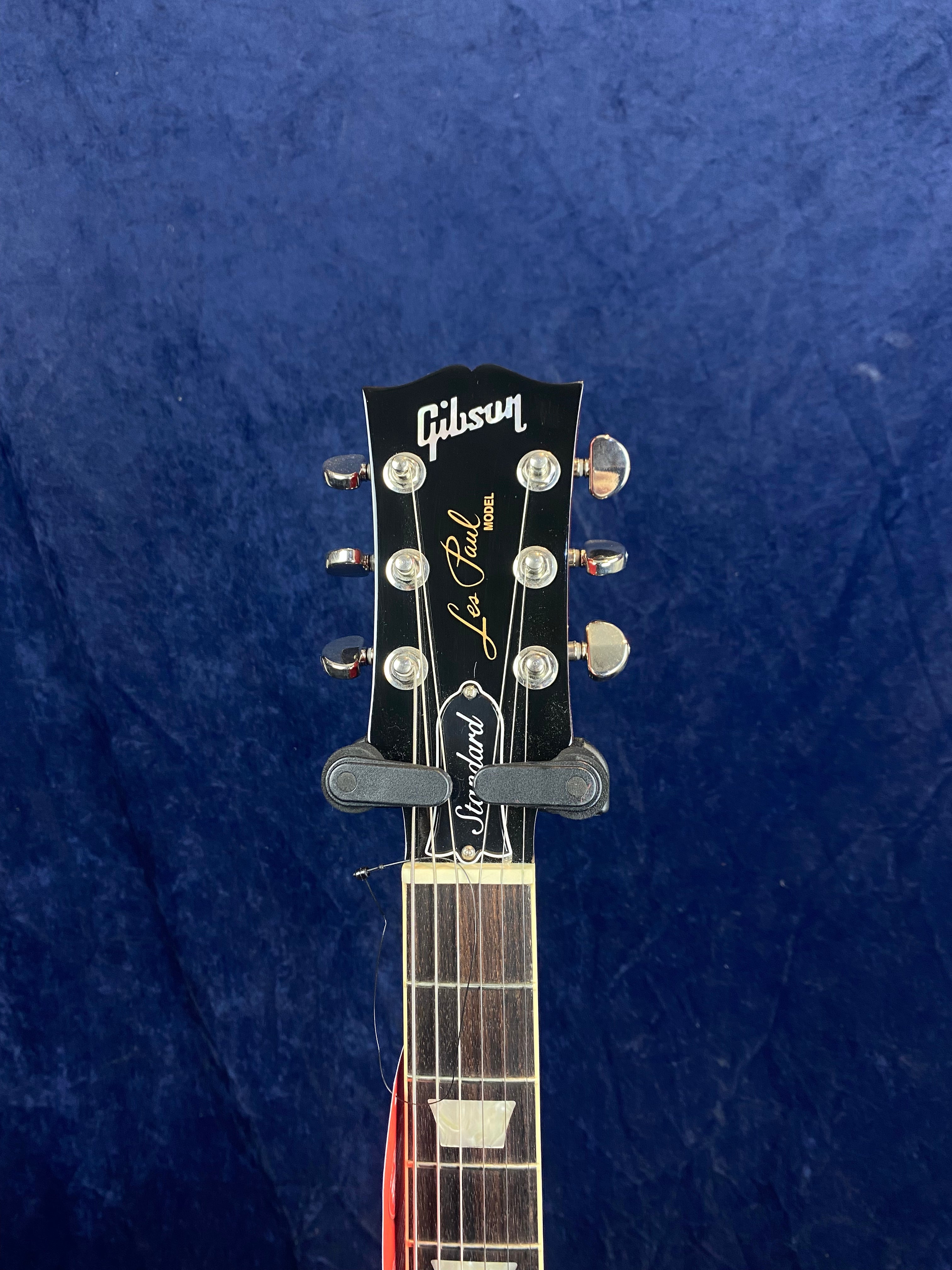 Headstock of a Gibson Les Paul electric guitar with six tuning pegs, "Gibson" logo, and "Standard" truss rod cover against a dark blue background.