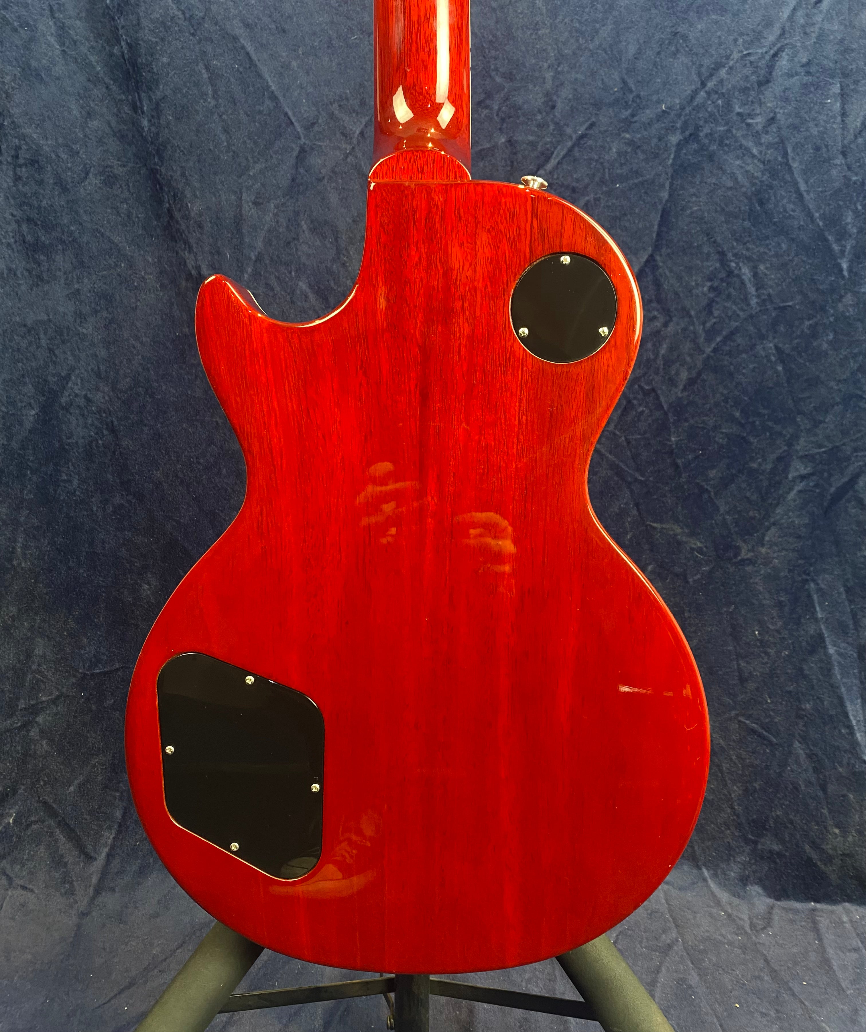 The back of a red electric guitar with a glossy finish, showing two black plastic covers for electronics access against a dark textured background.