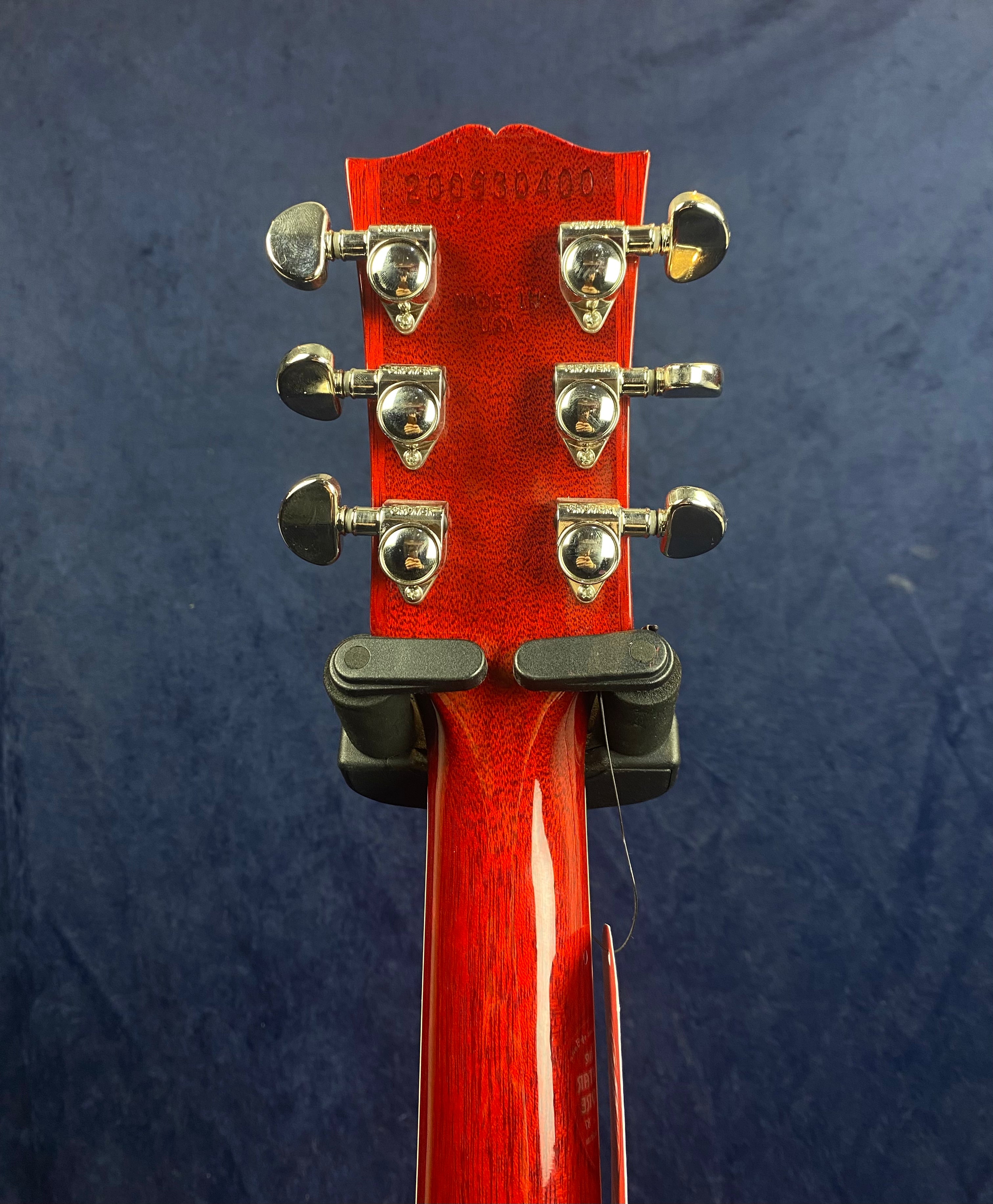 Back of a red electric guitar headstock showing six chrome tuning pegs, serial number, and “Made in USA” text, against a dark blue background.