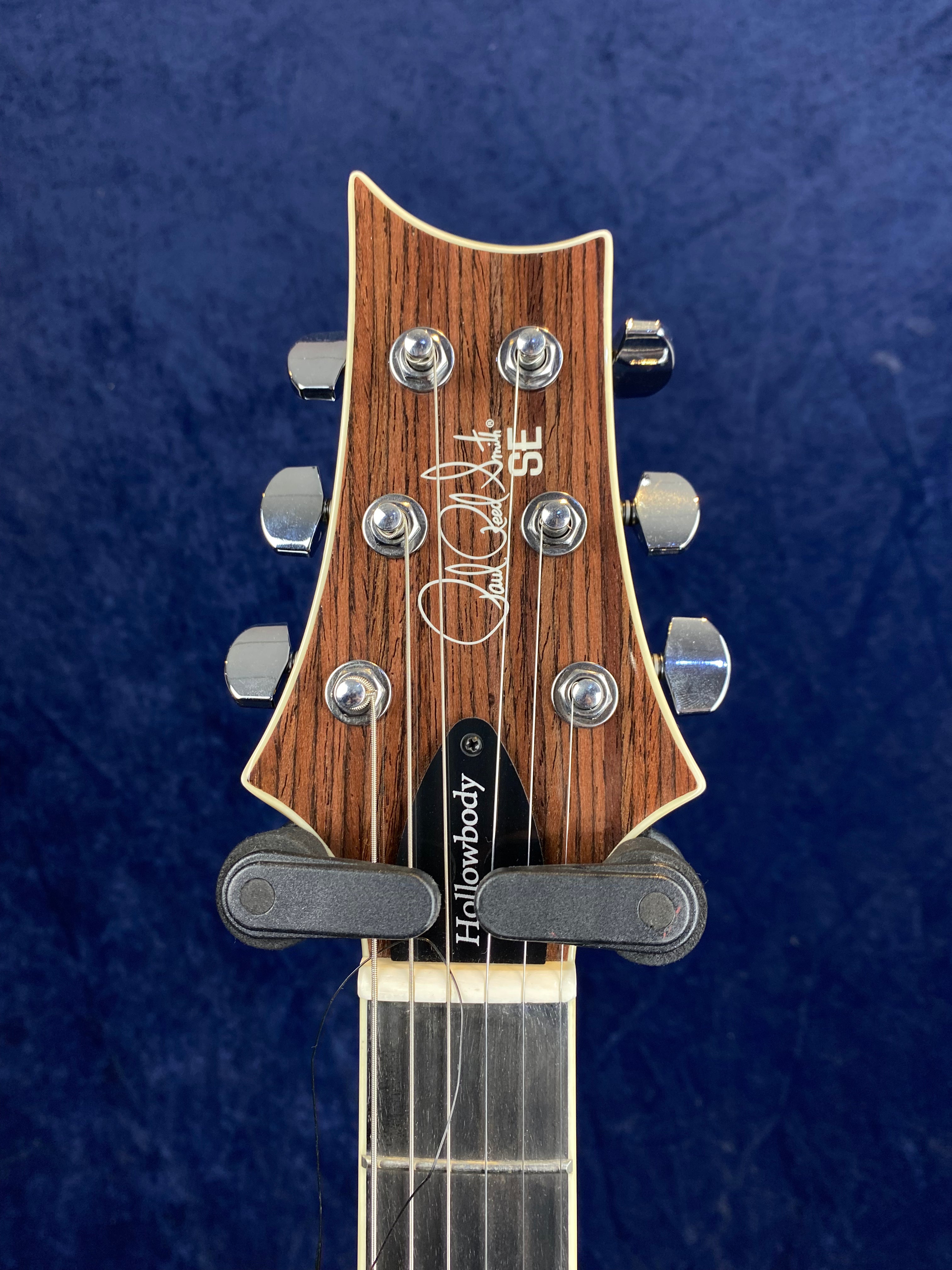 Electric guitar headstock with six chrome tuning pegs, wood grain finish, "SE" logo, and "Hollowbody" truss rod cover against a blue background.