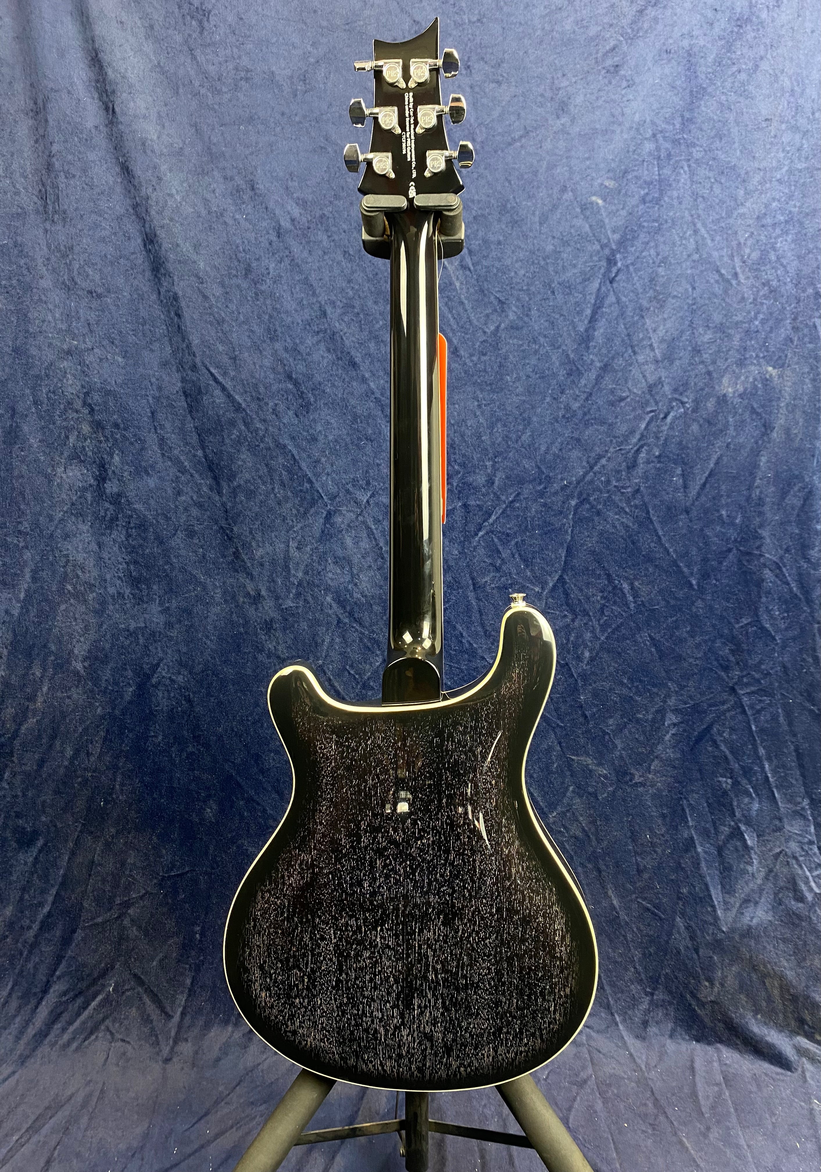 Back view of an electric guitar with a dark glossy finish, white body binding, and chrome tuning pegs, set against a dark blue textured background.