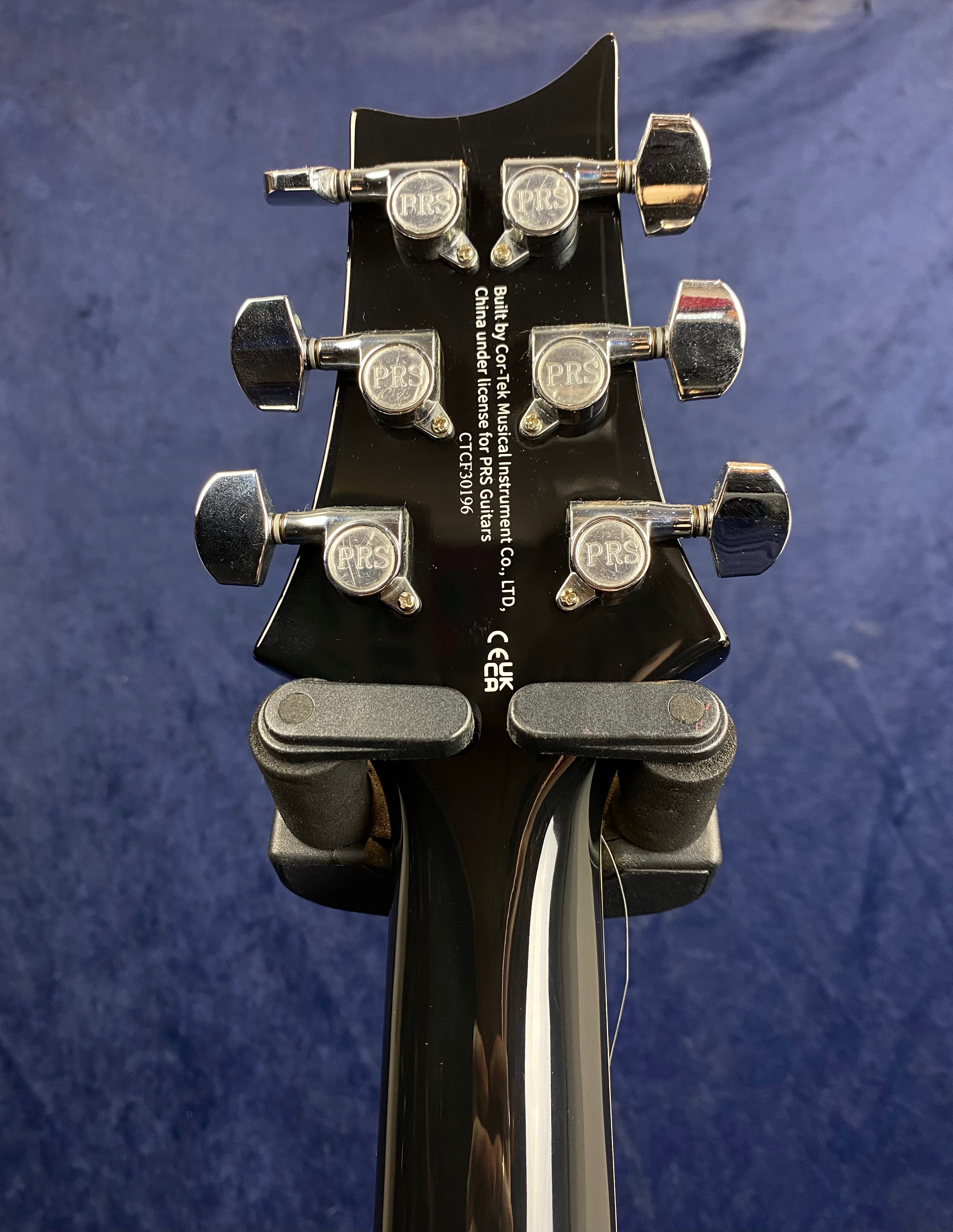 Back of a black electric guitar headstock with six PRS-branded chrome tuning pegs and manufacturer text in white.