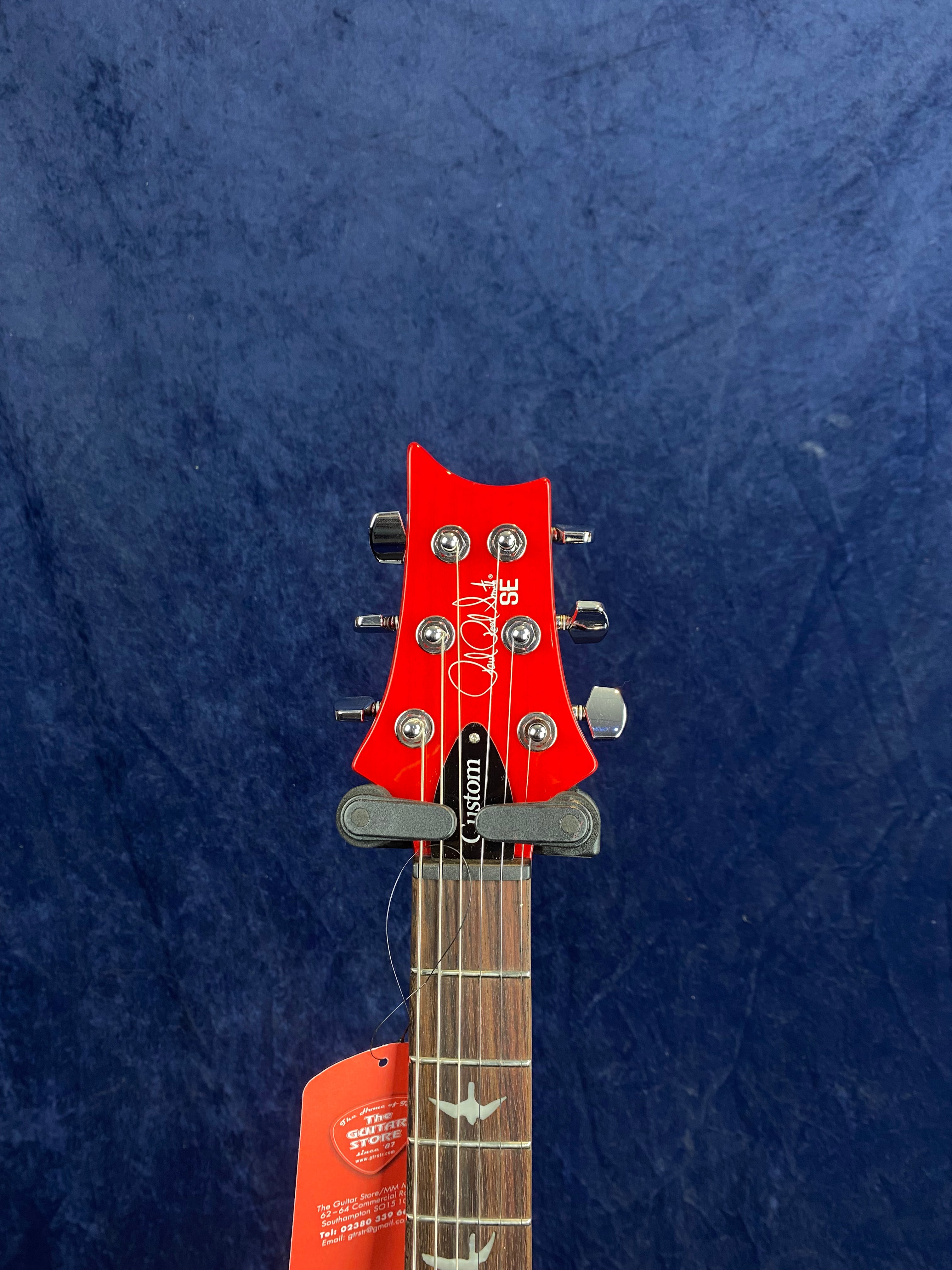Red headstock of a PRS SE electric guitar, chrome tuning pegs, rosewood fretboard with bird inlays, and a tag hanging from a string.