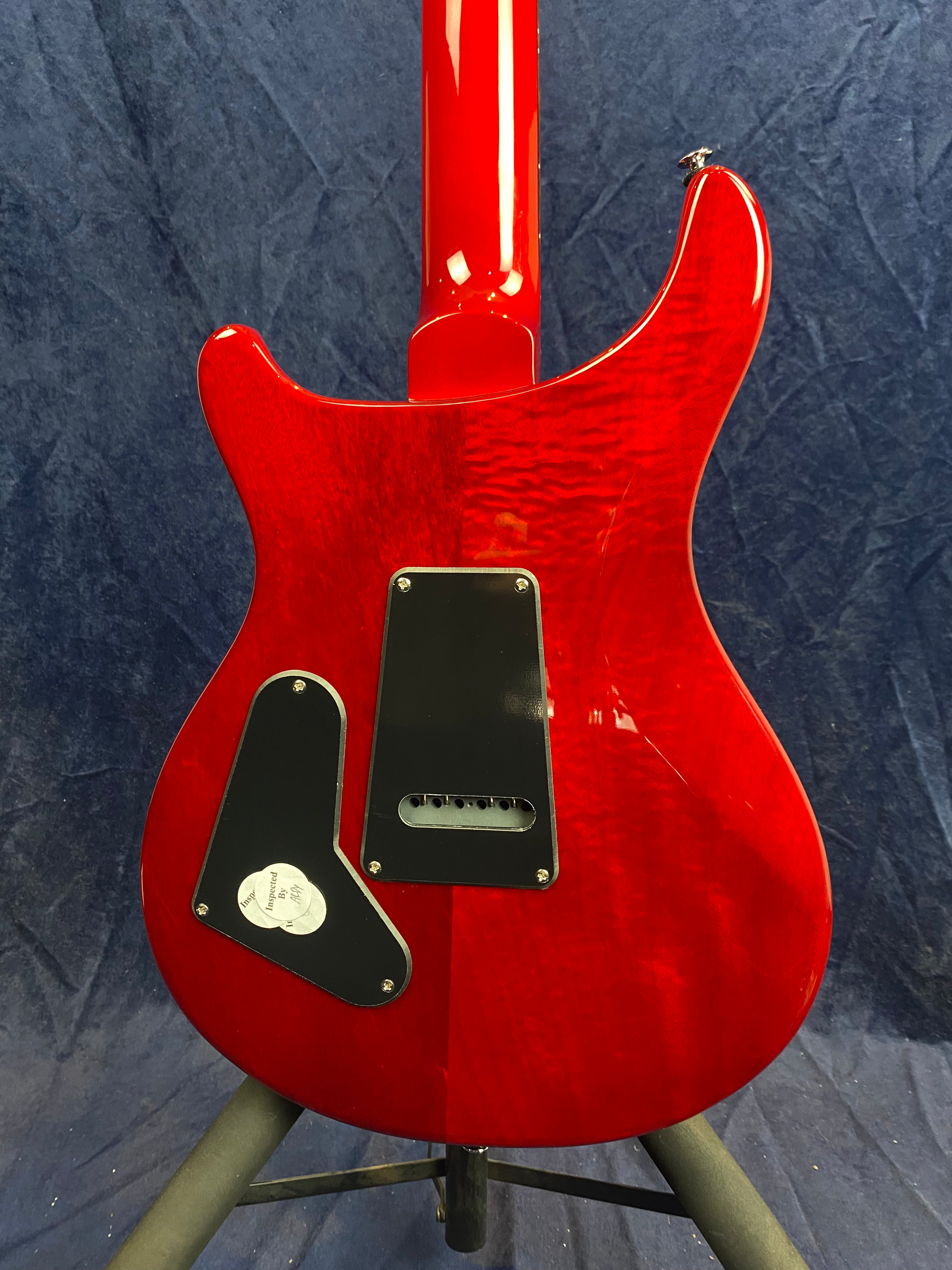 Glossy red electric guitar back shown with a curved body, two black plastic access panels, and mounted on a stand against a blue background.
