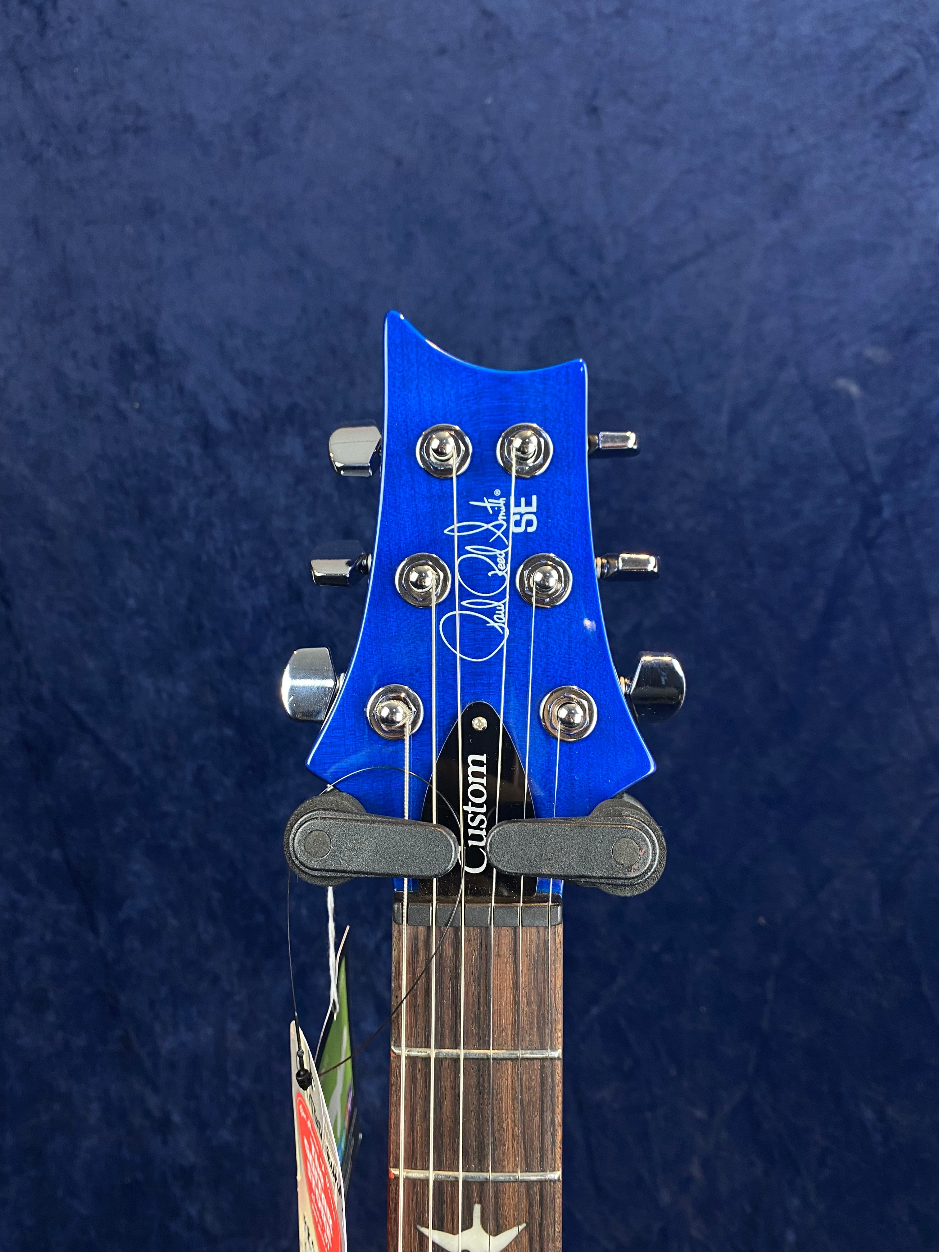 Blue electric guitar headstock with six chrome tuning pegs, white script logo, "SE" letters, "Custom" truss rod cover, and a bird inlay on the fretboard.