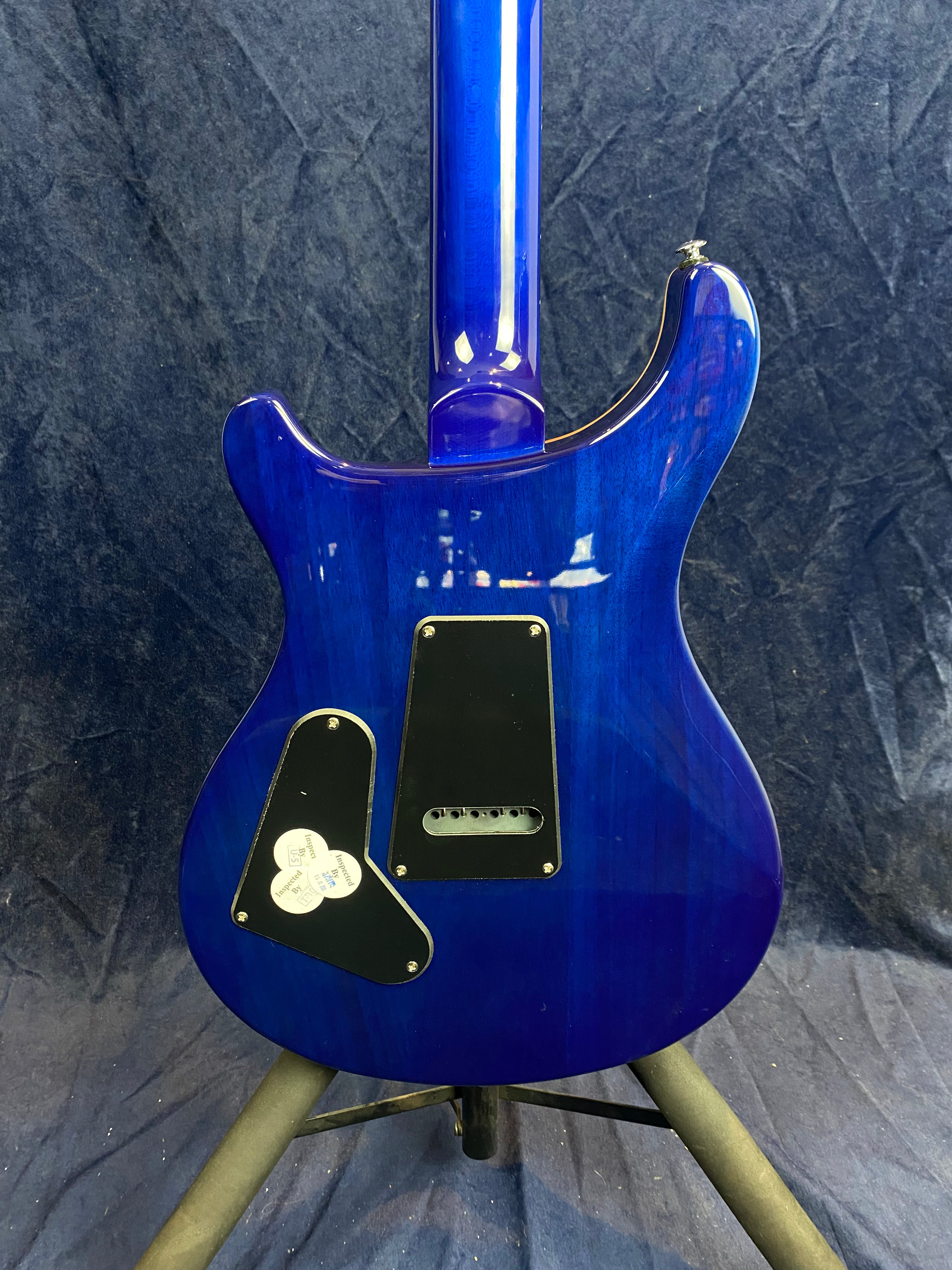 Blue electric guitar shown from the back, with two black plastic plates for electronics access and string holes visible on the body, standing on a tripod.