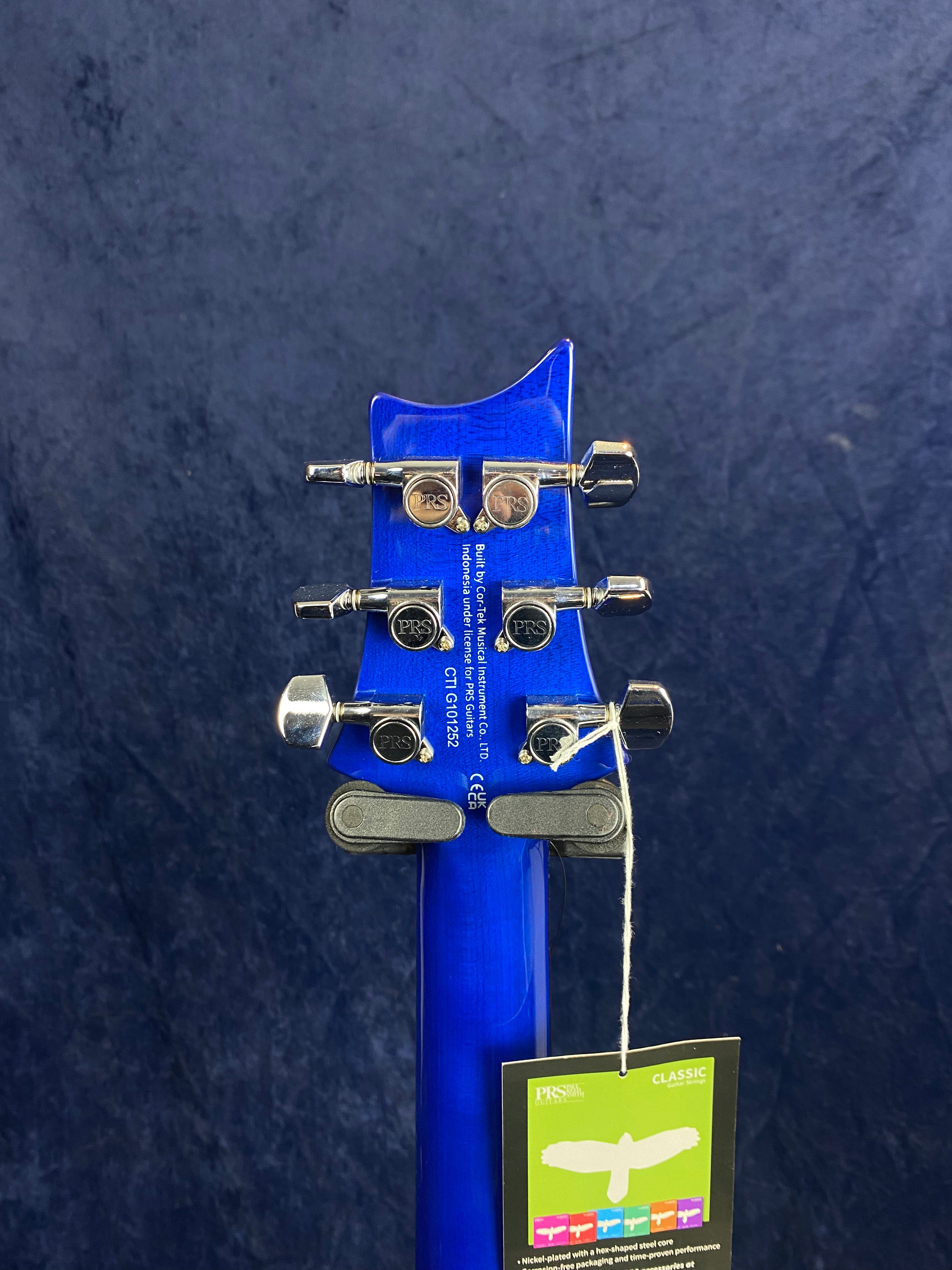 Blue electric guitar headstock with six silver tuning pegs, PRS logo visible, and a hanging product tag attached by string.