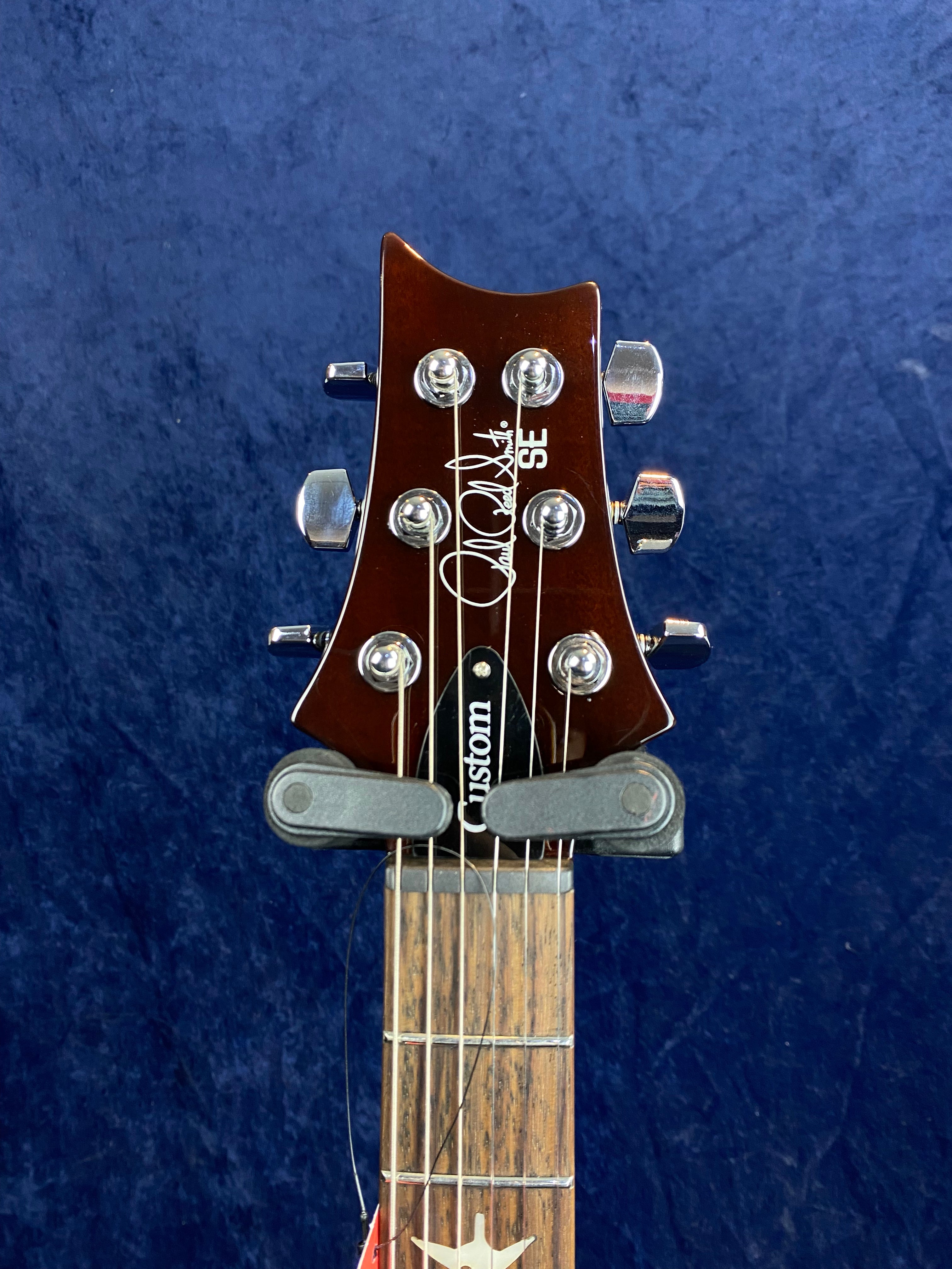 Brown PRS SE electric guitar headstock with chrome tuning pegs, “Custom” truss rod cover, and white signature on the front against a blue background.