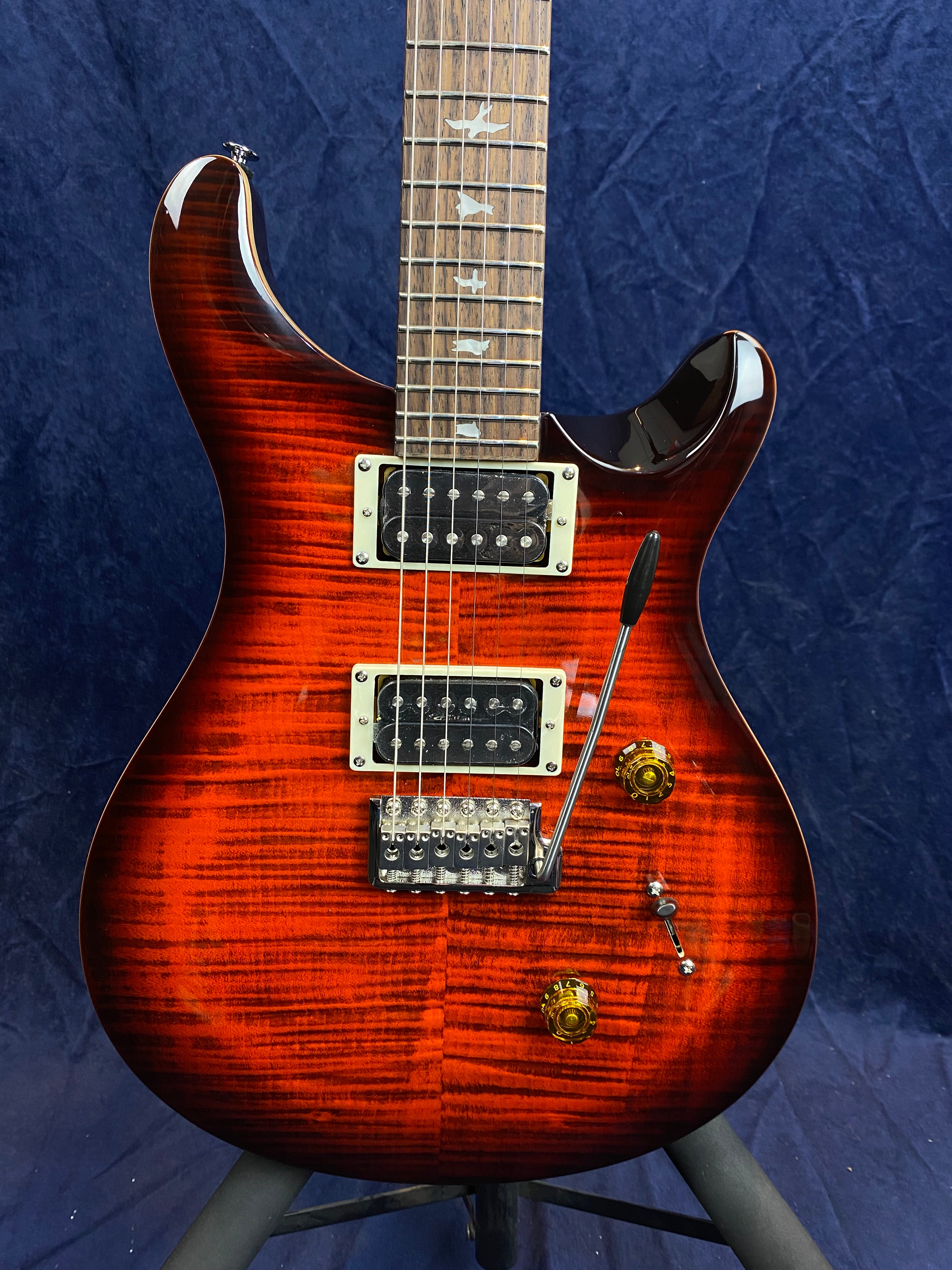 Electric guitar with a glossy red-orange flamed wood finish, two humbucker pickups, gold control knobs, whammy bar, and bird inlays on the fretboard.