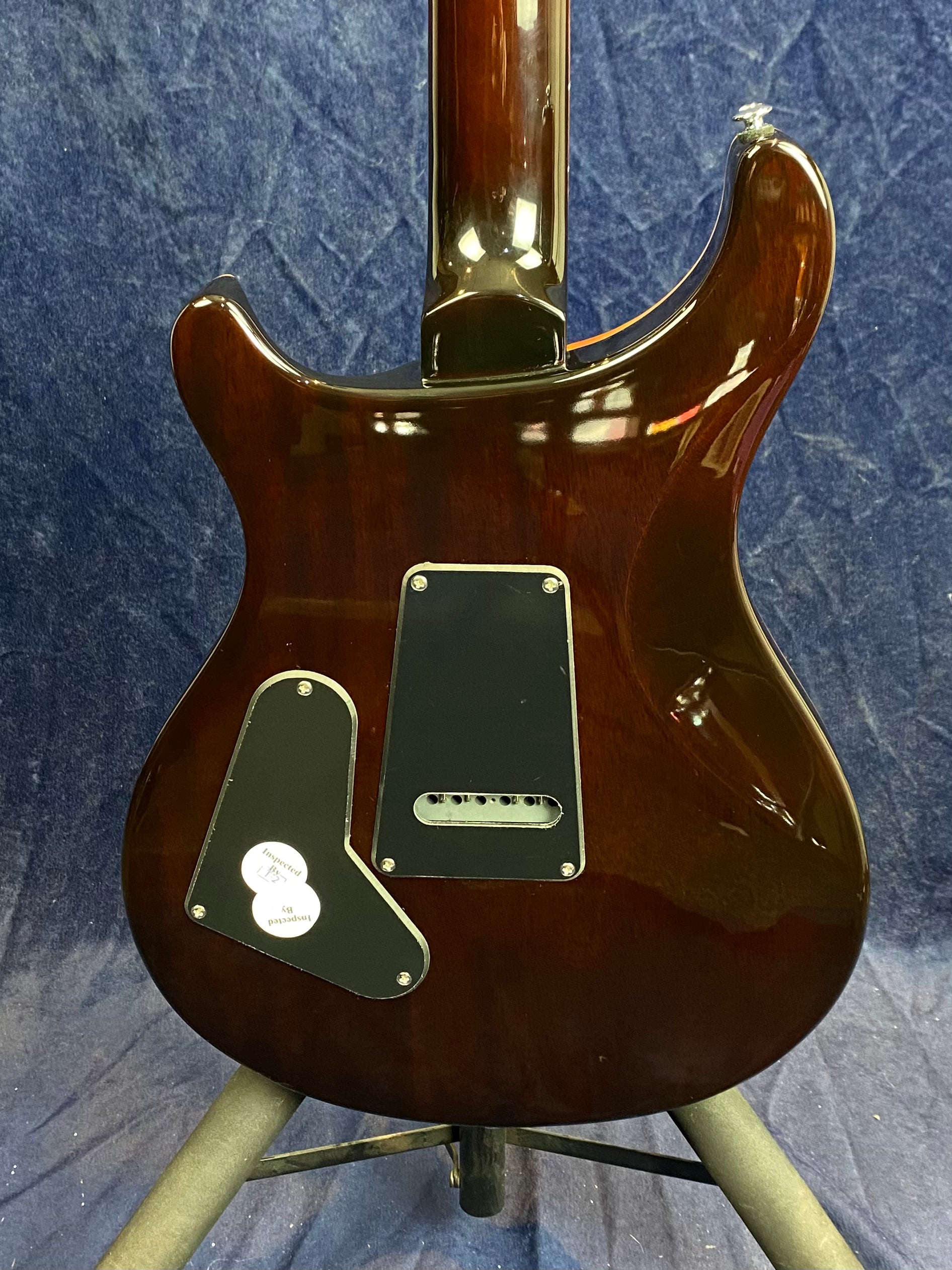 The back of a glossy brown electric guitar with two black plastic covers and an "Inspected" sticker is shown upright on a stand.