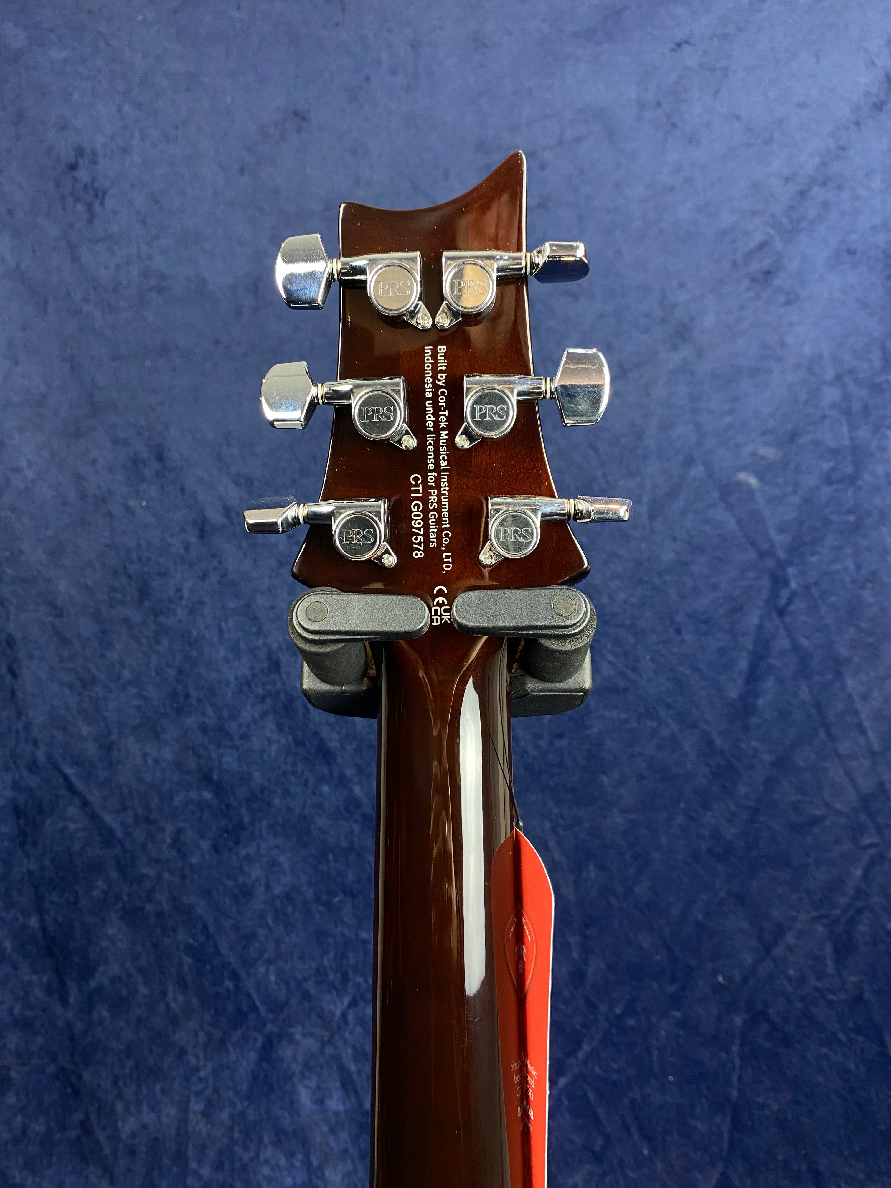 The back of an electric guitar headstock with six chrome PRS tuning pegs, wood finish neck, and white manufacturing text visible at the center.