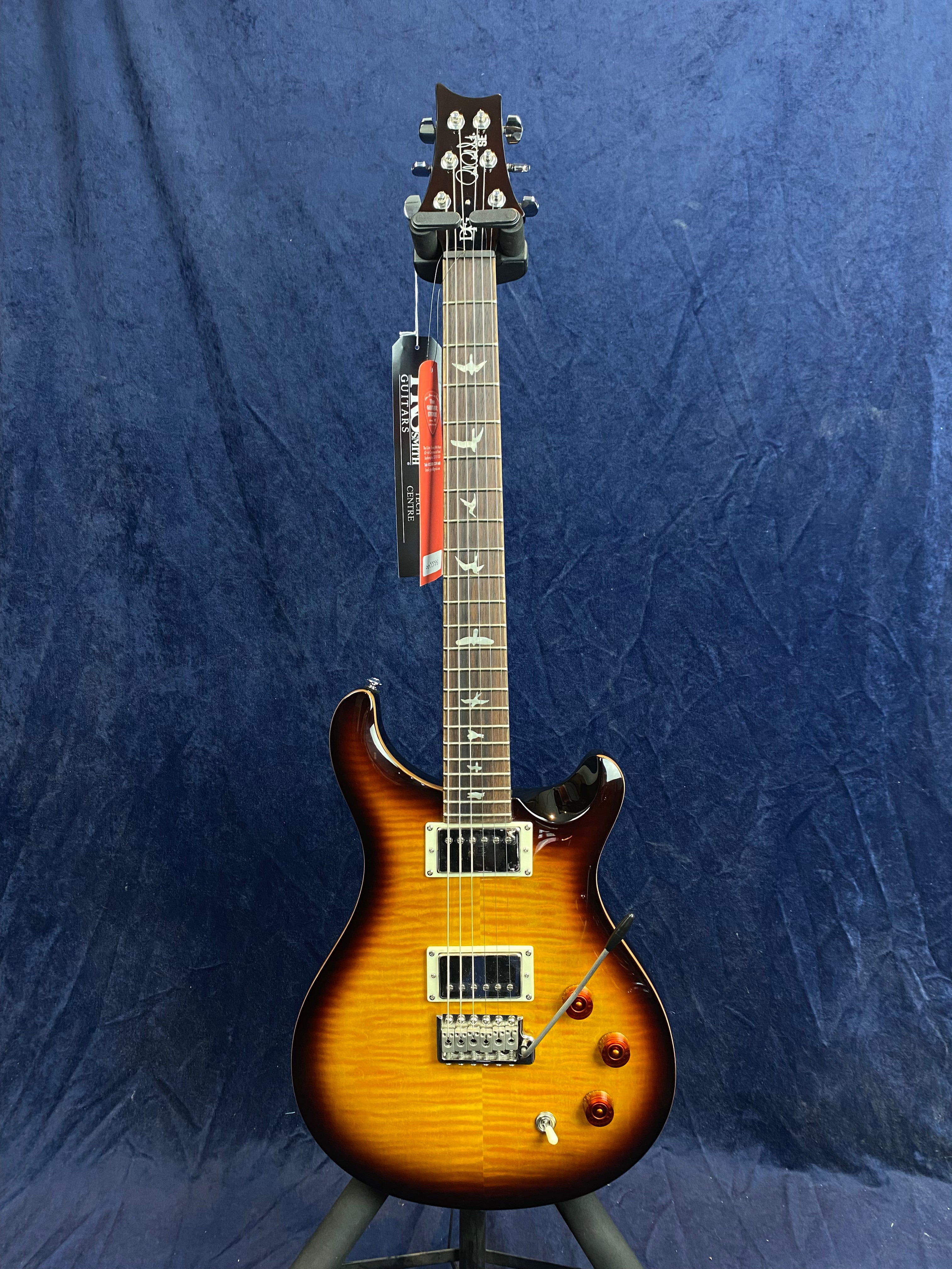 Sunburst electric guitar with dual humbucker pickups, bird inlays on the fretboard, chrome hardware, and three amber control knobs on a dark blue backdrop.