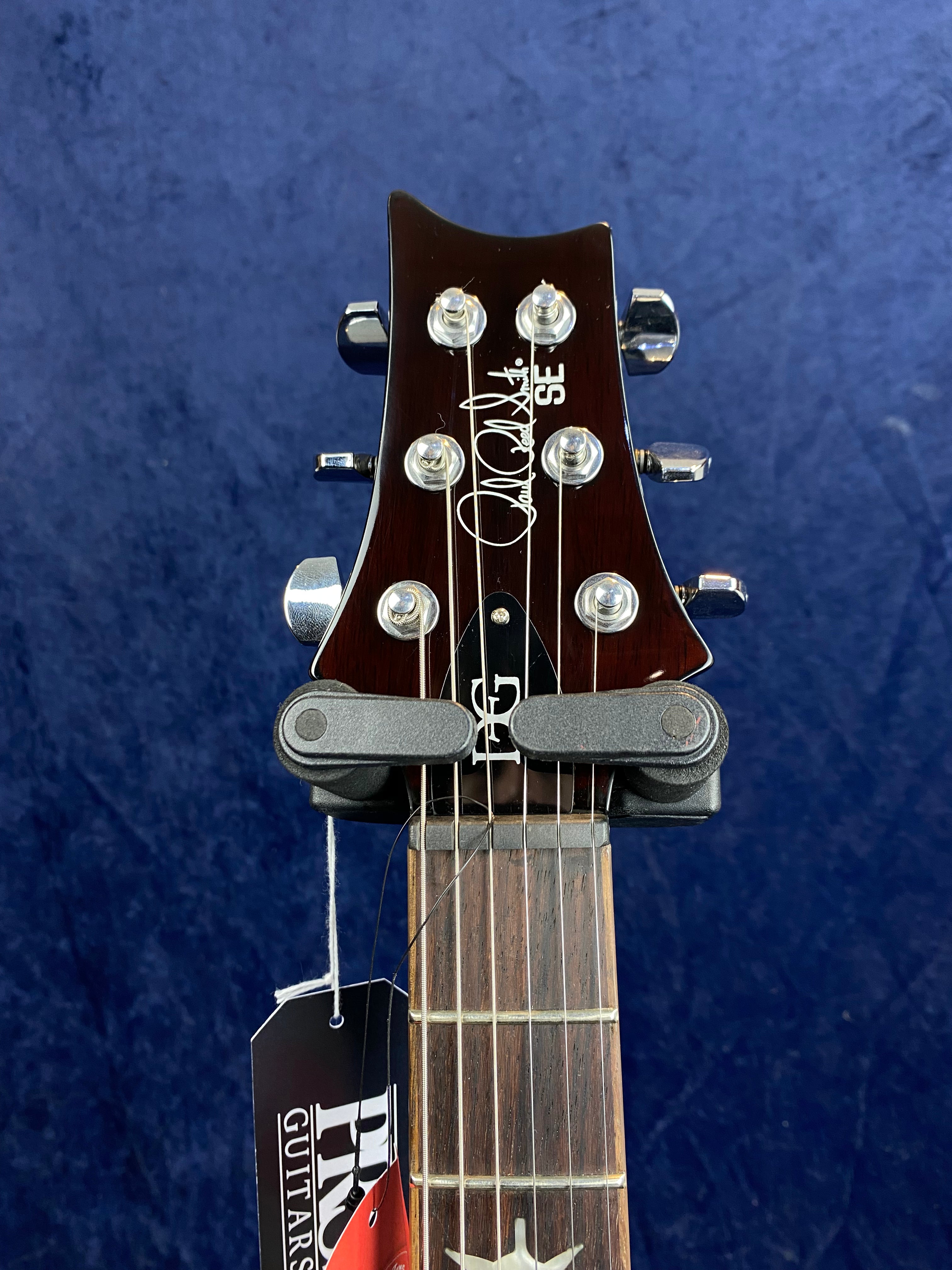 Headstock of a PRS SE electric guitar with chrome tuning pegs, signature logo, string tag attached, and a dark wood finish against a blue background.