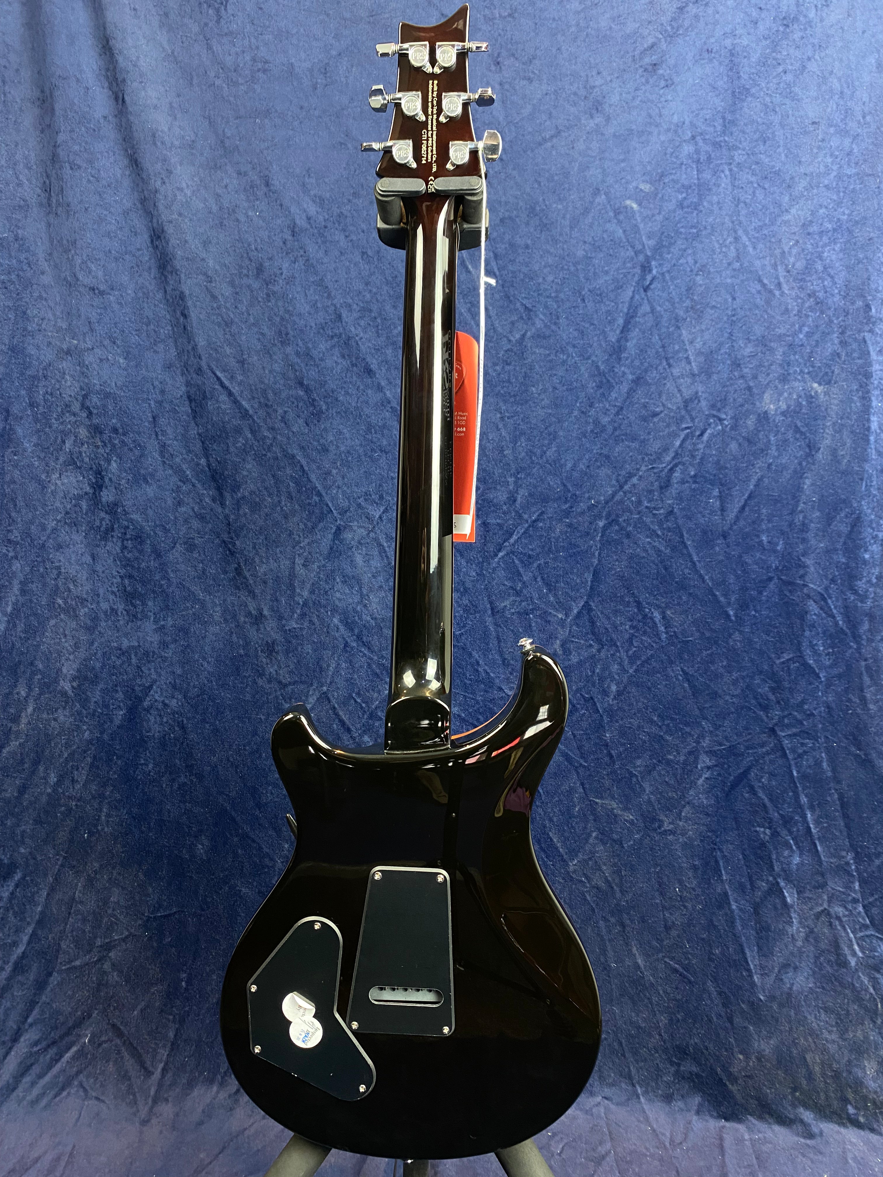 Glossy black electric guitar shown from the back, revealing metal rear covers and tuning pegs, against a dark blue textured background.