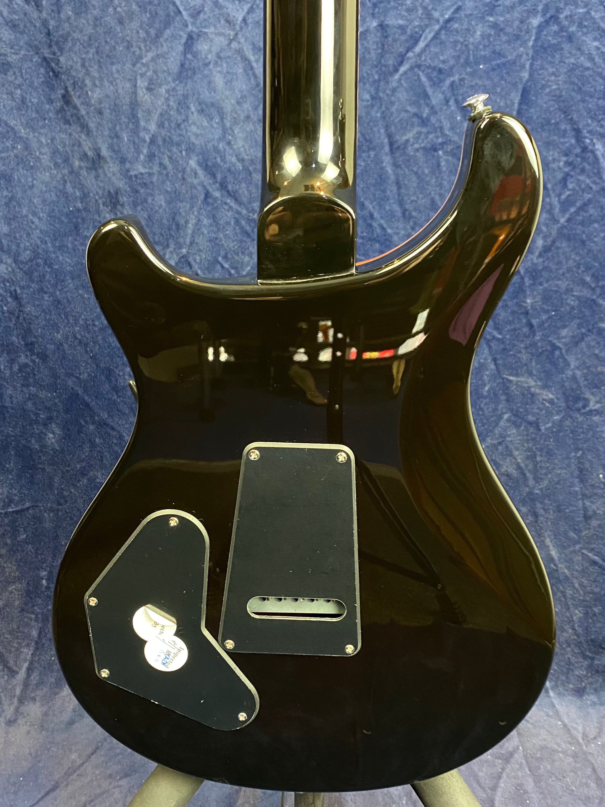 Glossy black electric guitar back, two control cavity covers held by screws, neck joint at top center, blue background.