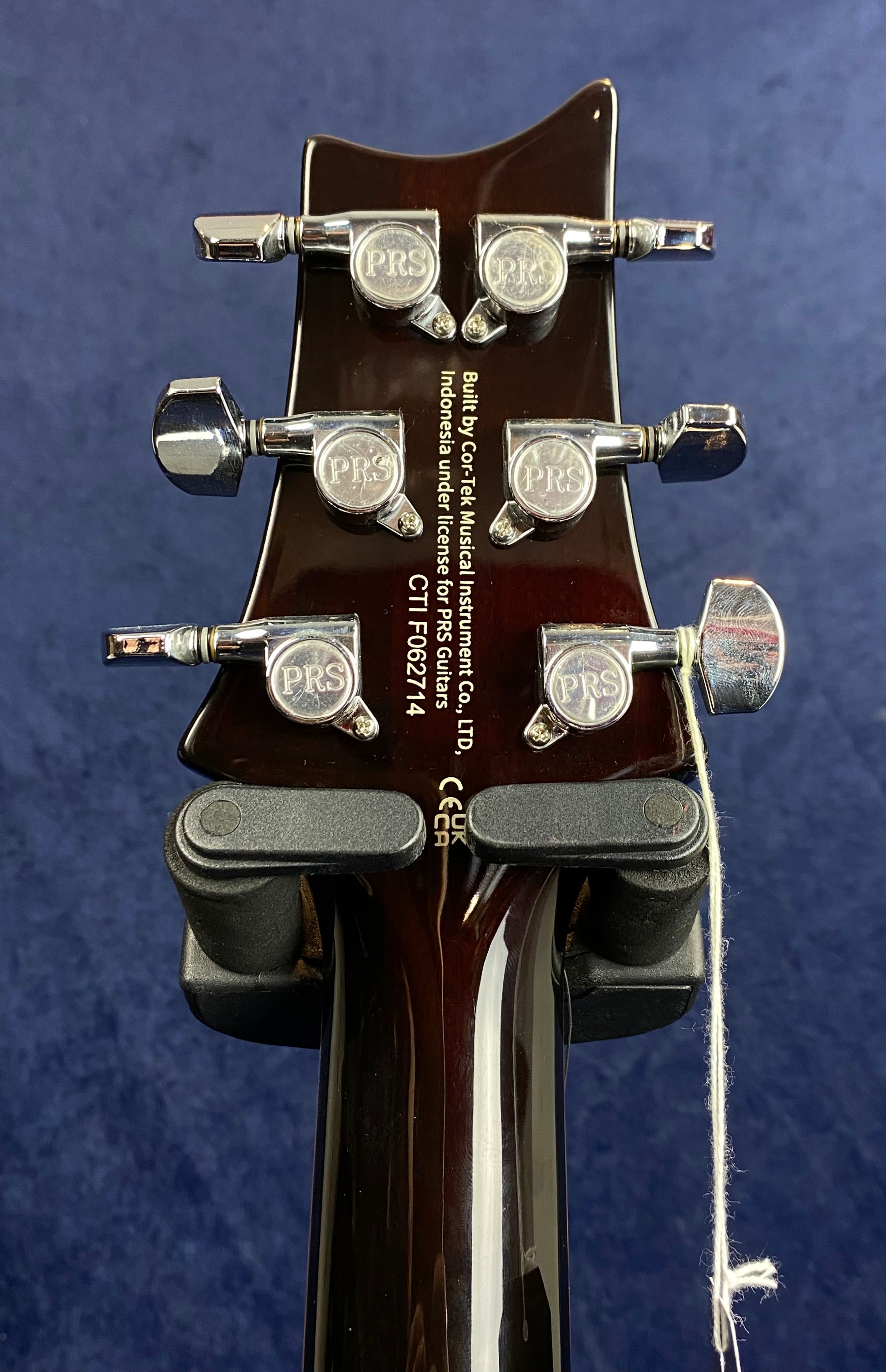 Six chrome tuning pegs with "PRS" logos on a dark brown electric guitar headstock, serial number CTIF062714, text indicating made in Indonesia.