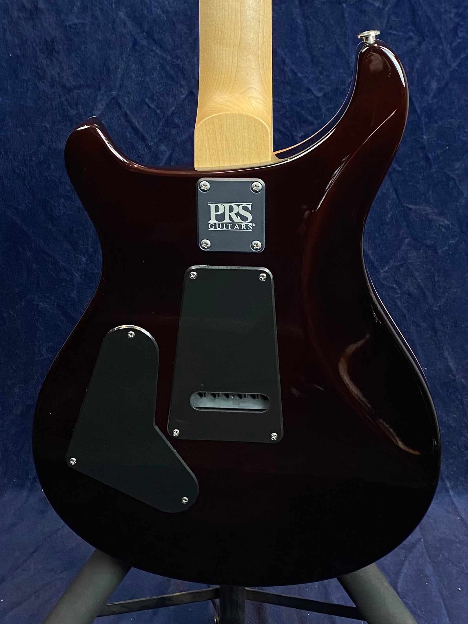 Back view of a PRS electric guitar showing a wooden neck, dark glossy body, and two black plastic cover plates with a visible screw-on neck plate.