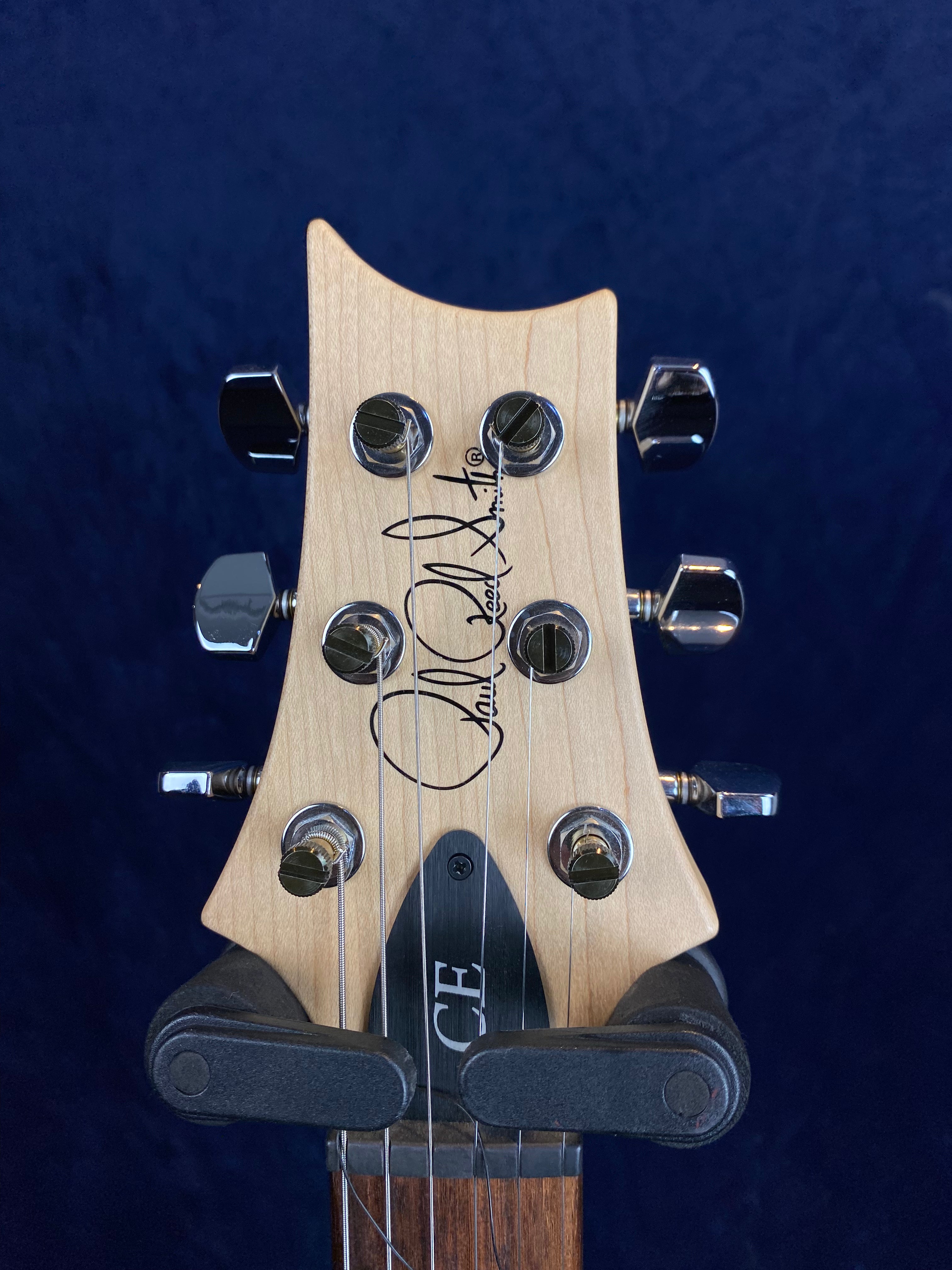 Headstock of an electric guitar with six chrome tuning pegs, wood finish, signature logo, and “CE” label on a black truss rod cover.