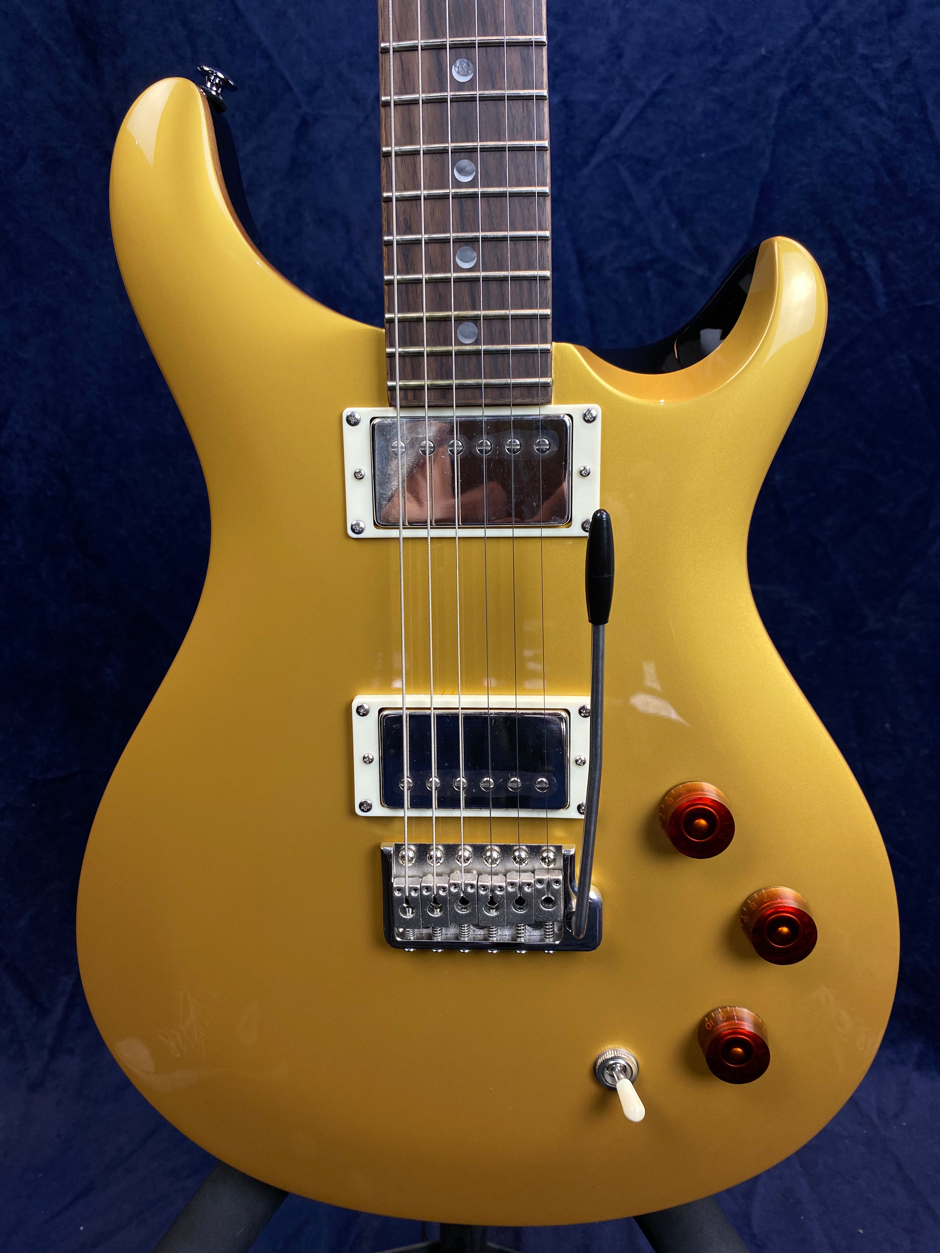 Gold electric guitar with two chrome pickups, three amber control knobs, a whammy bar, and a toggle switch on a dark blue background.