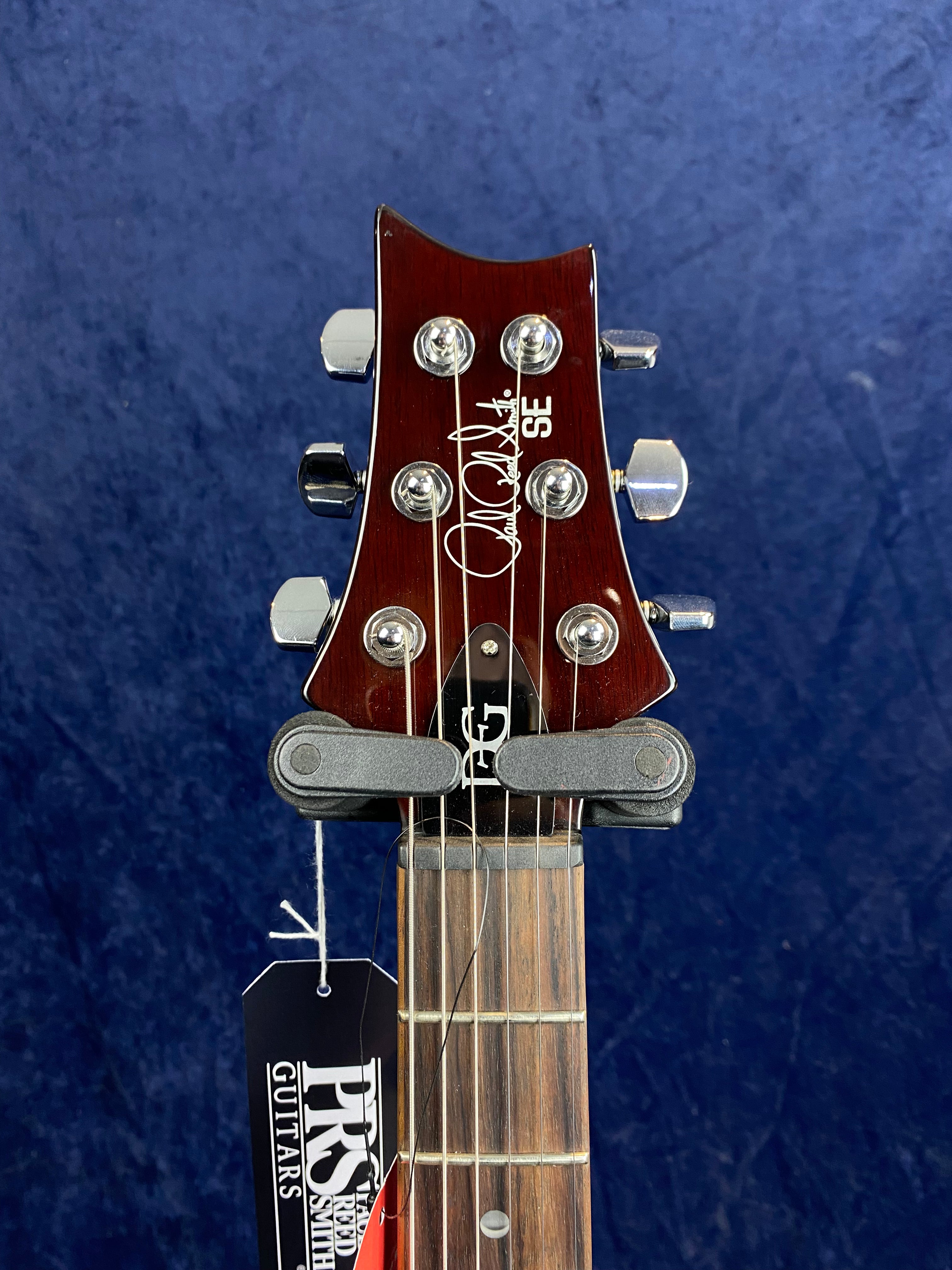 Close-up of a PRS SE electric guitar headstock with chrome tuners, visible logo, signature, and a hanging PRS Guitars tag against a dark blue background.