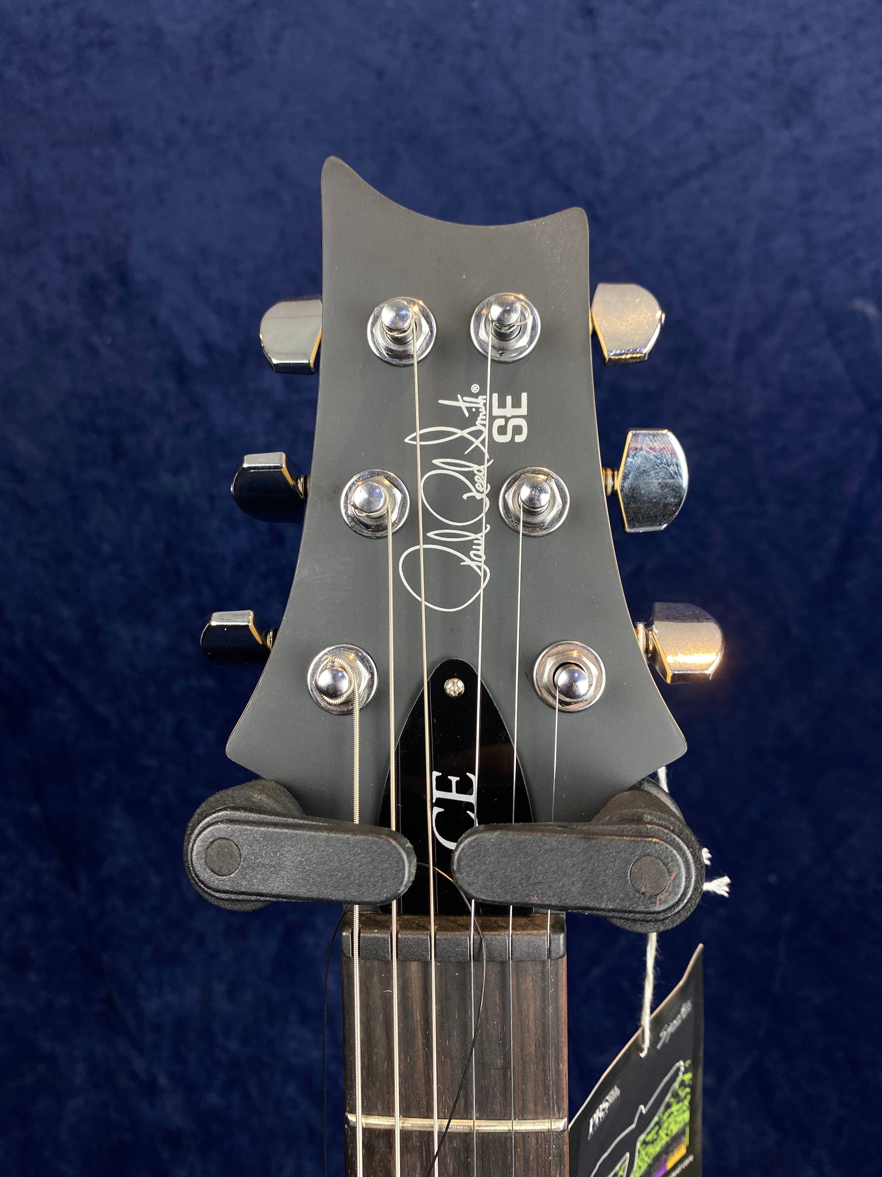 Electric guitar headstock with six chrome tuners, a black finish, and "SE" plus a signature logo in white script near the top.