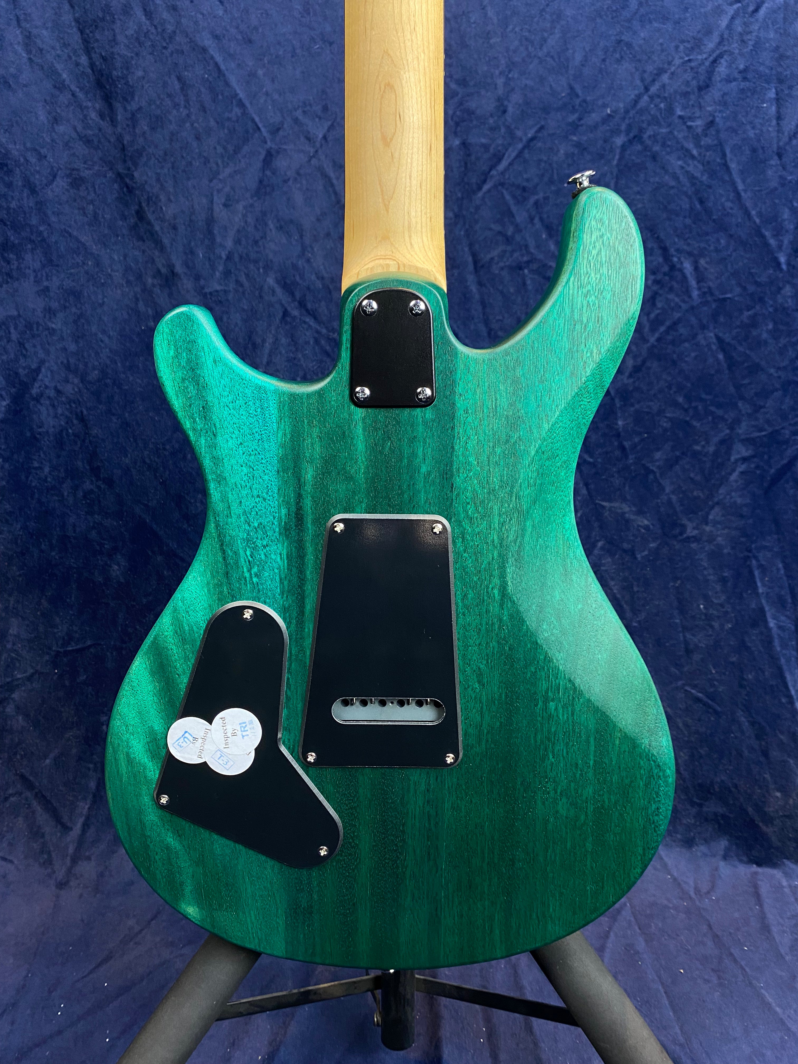 Electric guitar shown from the back, with a green stained wood body, black plastic backplates, and a natural wood neck against a dark blue background.