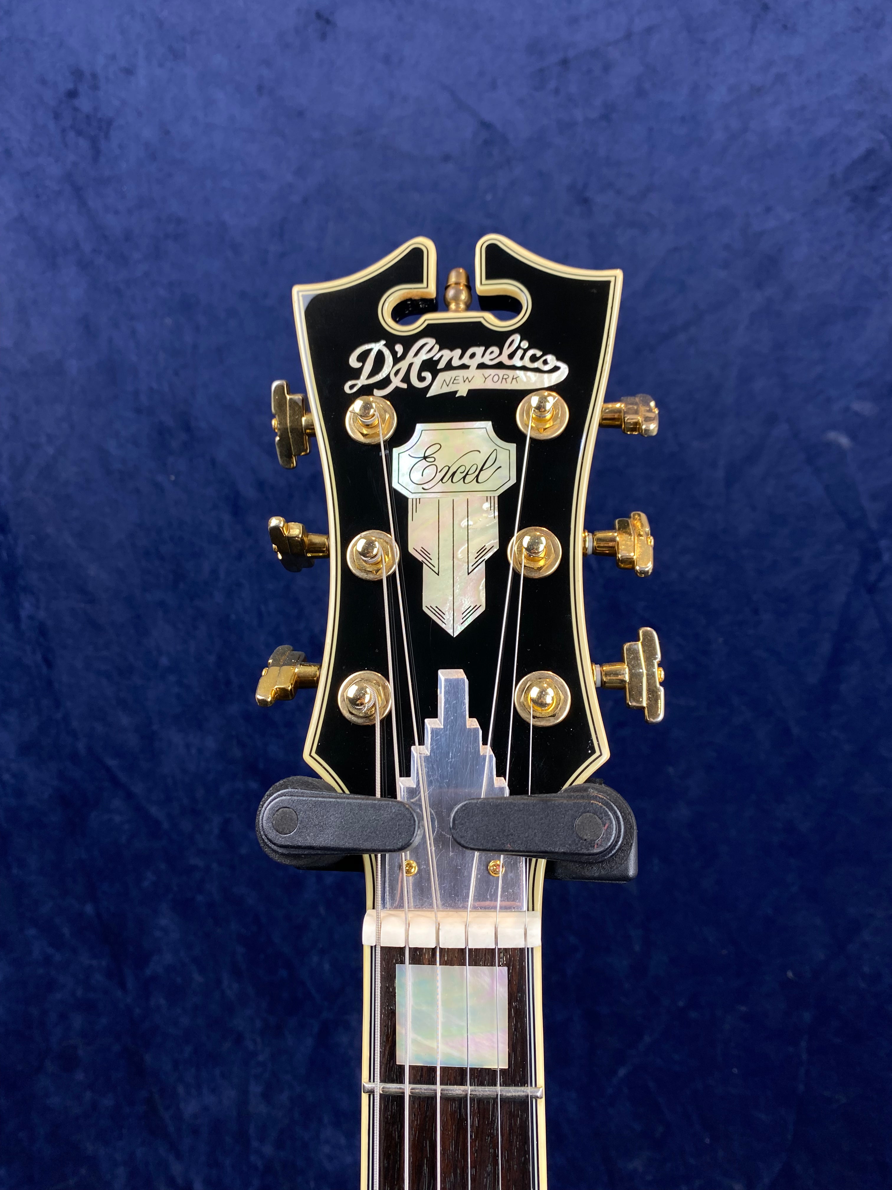 D'Angelico Excel electric guitar headstock with gold tuners, decorative inlays, and a black background with white and gold details.