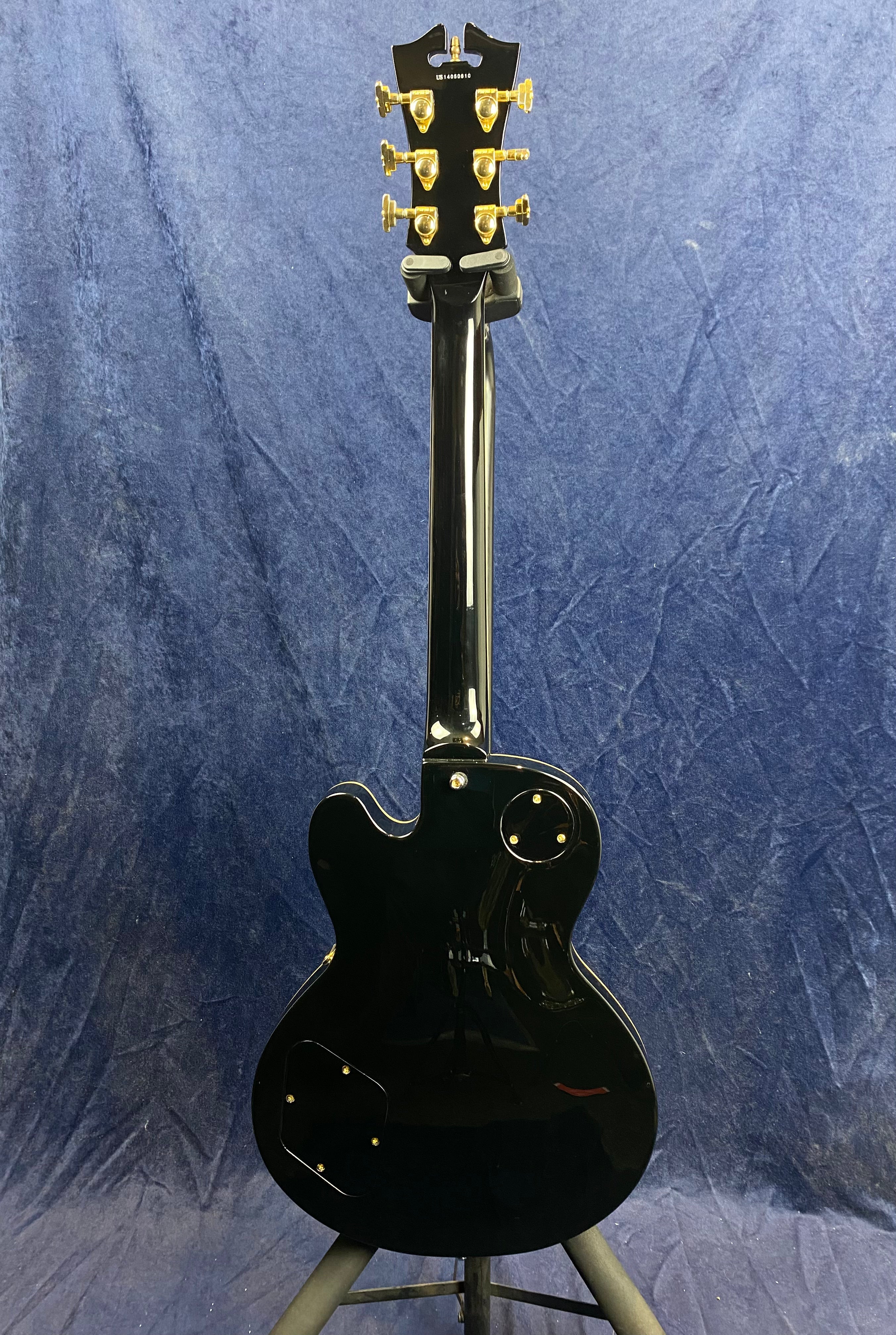 Glossy black electric guitar shown from the back, gold tuning pegs, single cutaway body, set on a stand against a blue backdrop.