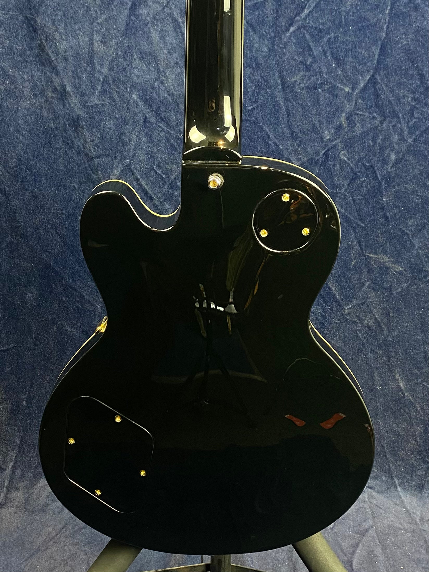 Glossy black electric guitar shown from the back, with two access panels secured by gold screws and a single cutaway body design.