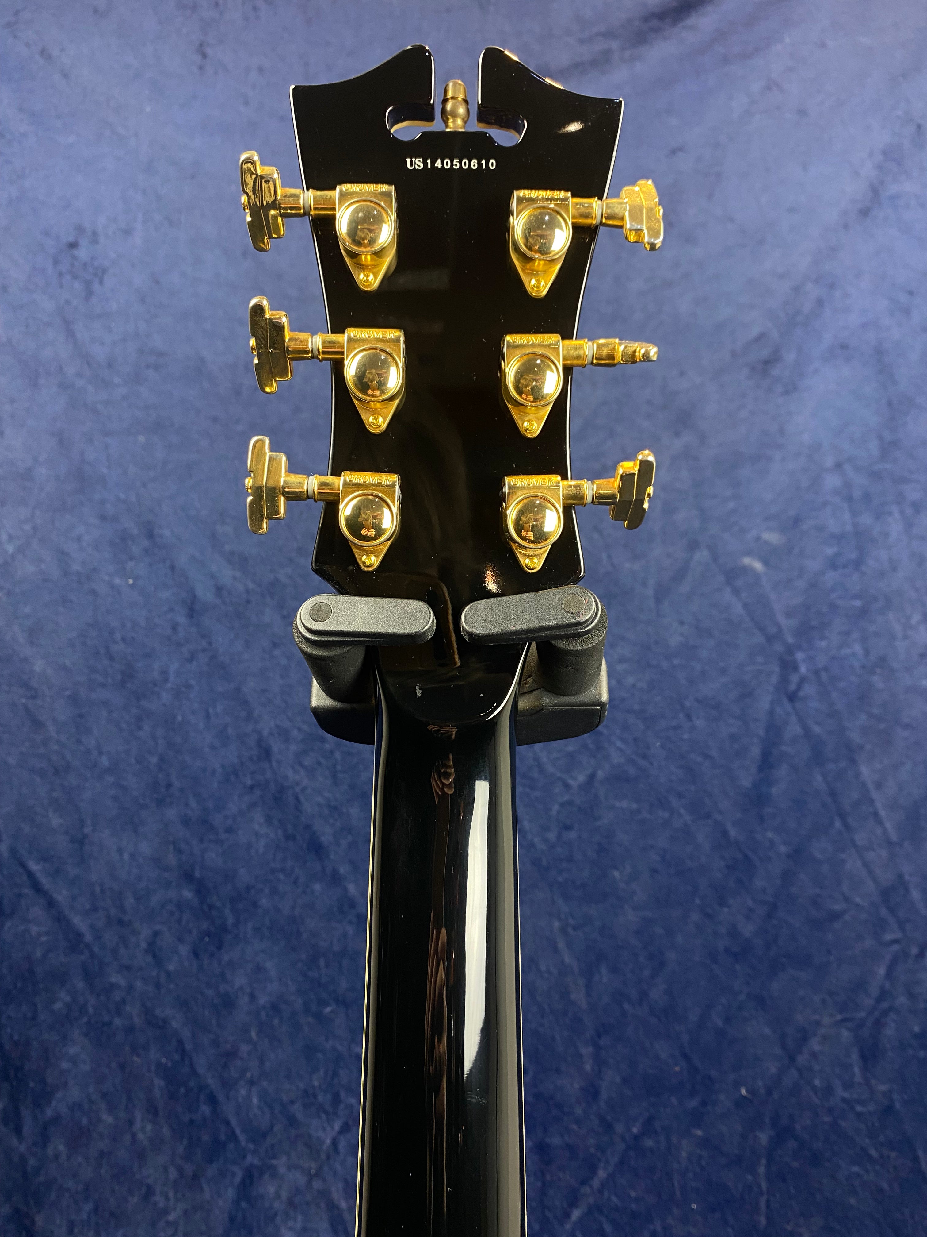 Back of an electric guitar headstock with six gold tuning pegs and a serial number US14050610 against a dark blue background.