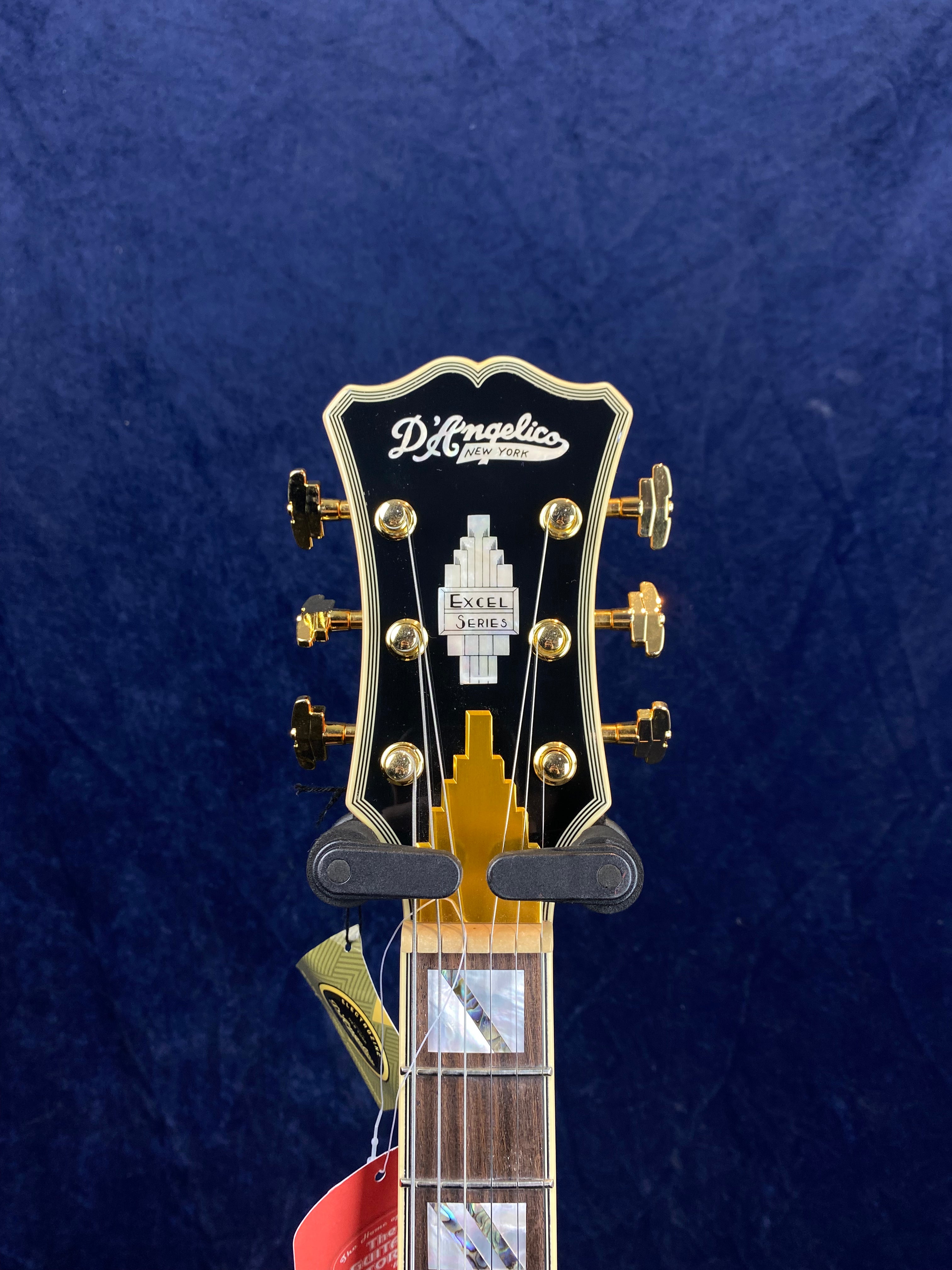Headstock of a D’Angelico New York electric guitar, gold tuners, art deco inlay labeled "Excel Series," strings attached, dark blue background.