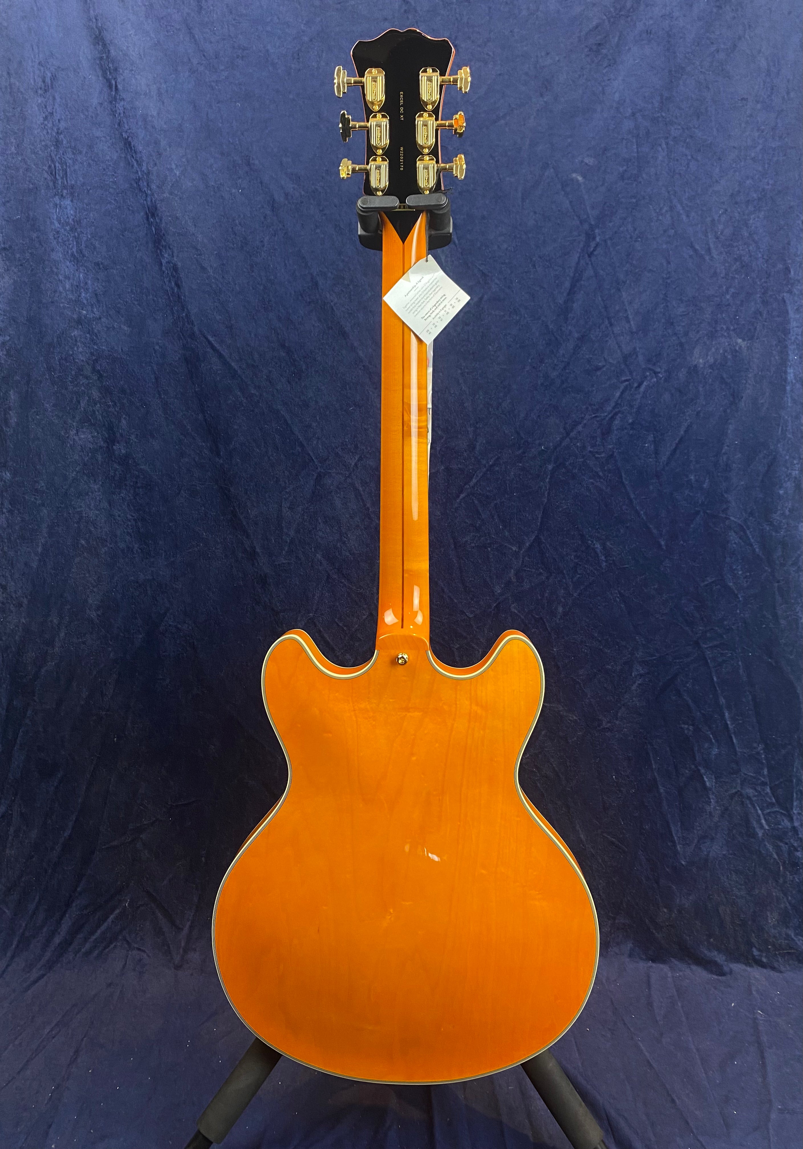 Orange semi-hollow electric guitar shown from the back, with gold tuning pegs and a tag attached to the neck against a dark blue background.