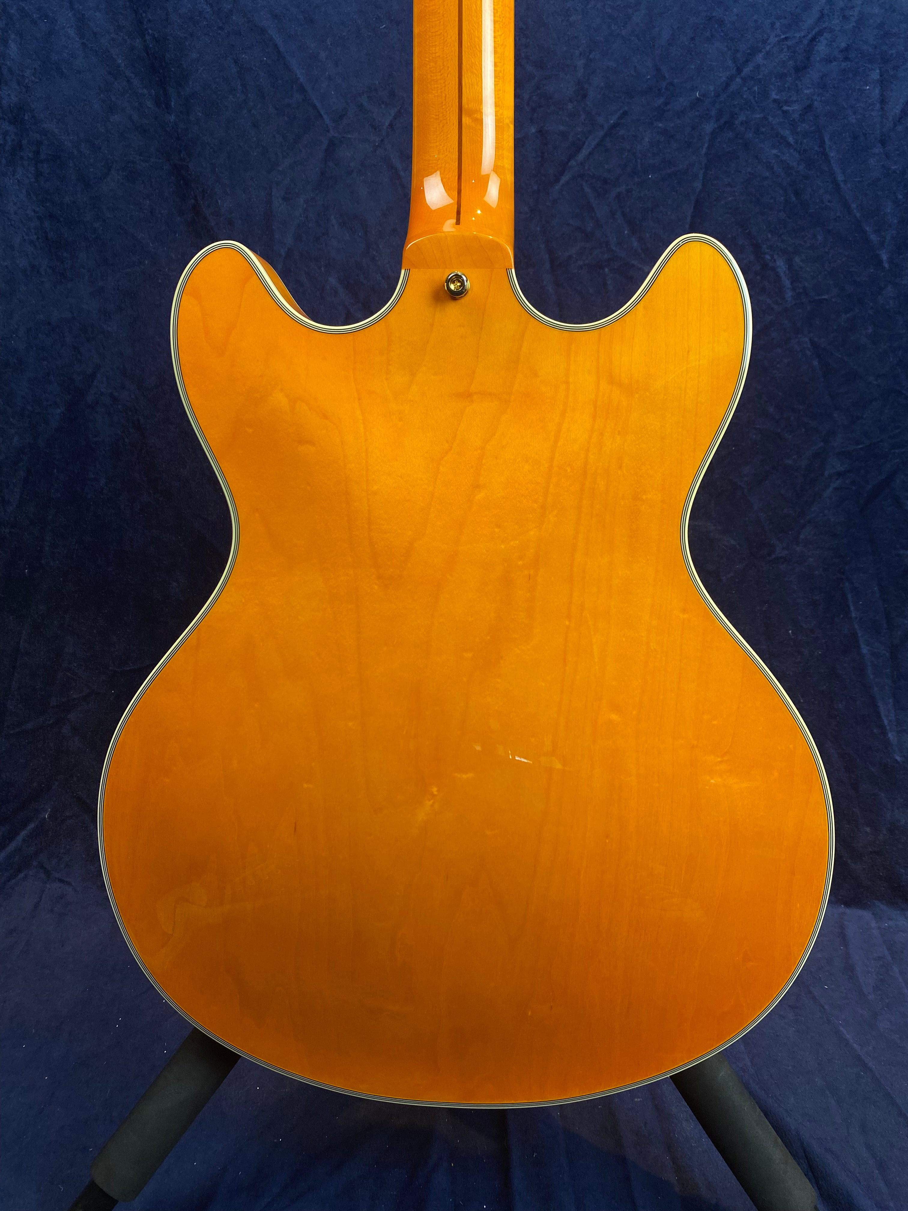 Amber-colored electric guitar back with a double cutaway shape, visible wood grain, cream binding, and a glossy finish against a dark blue background.