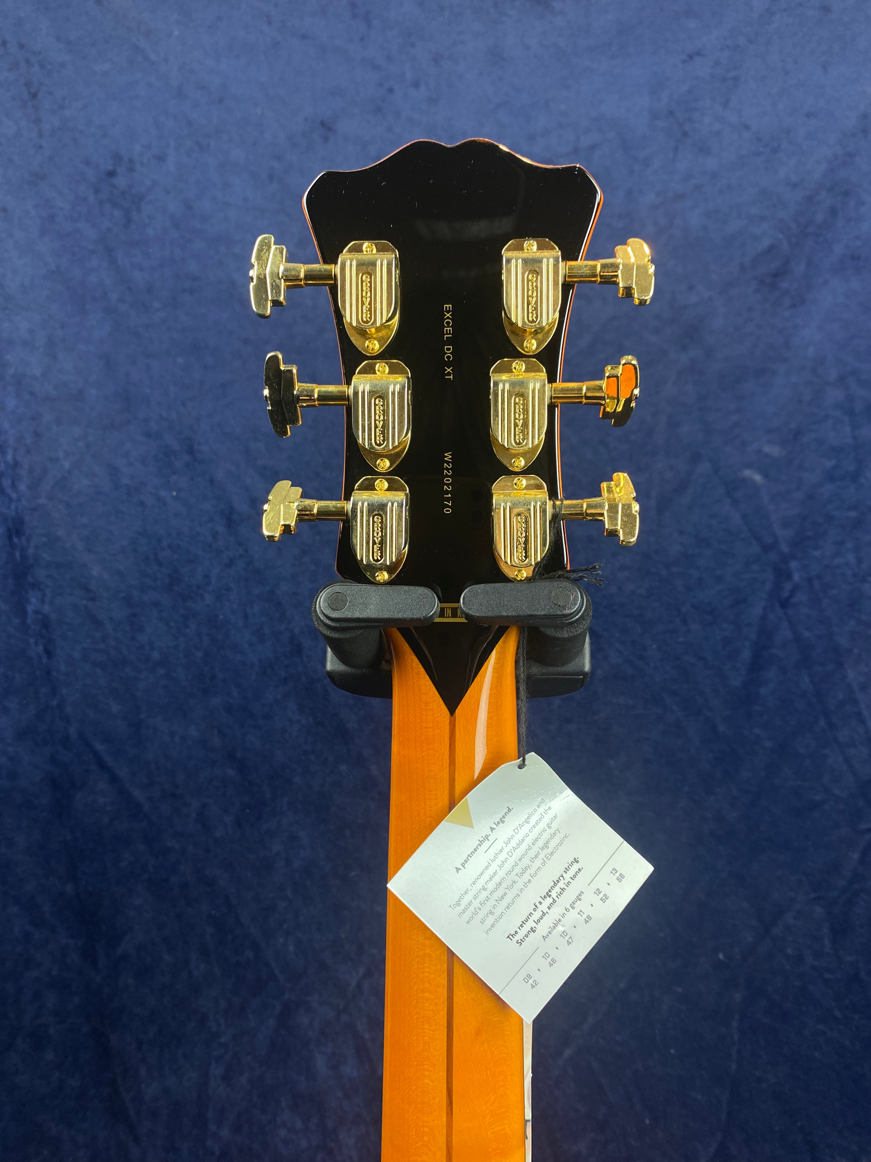 Back of electric guitar headstock with six gold tuning pegs, product label attached to the neck, and a dark blue textured background.