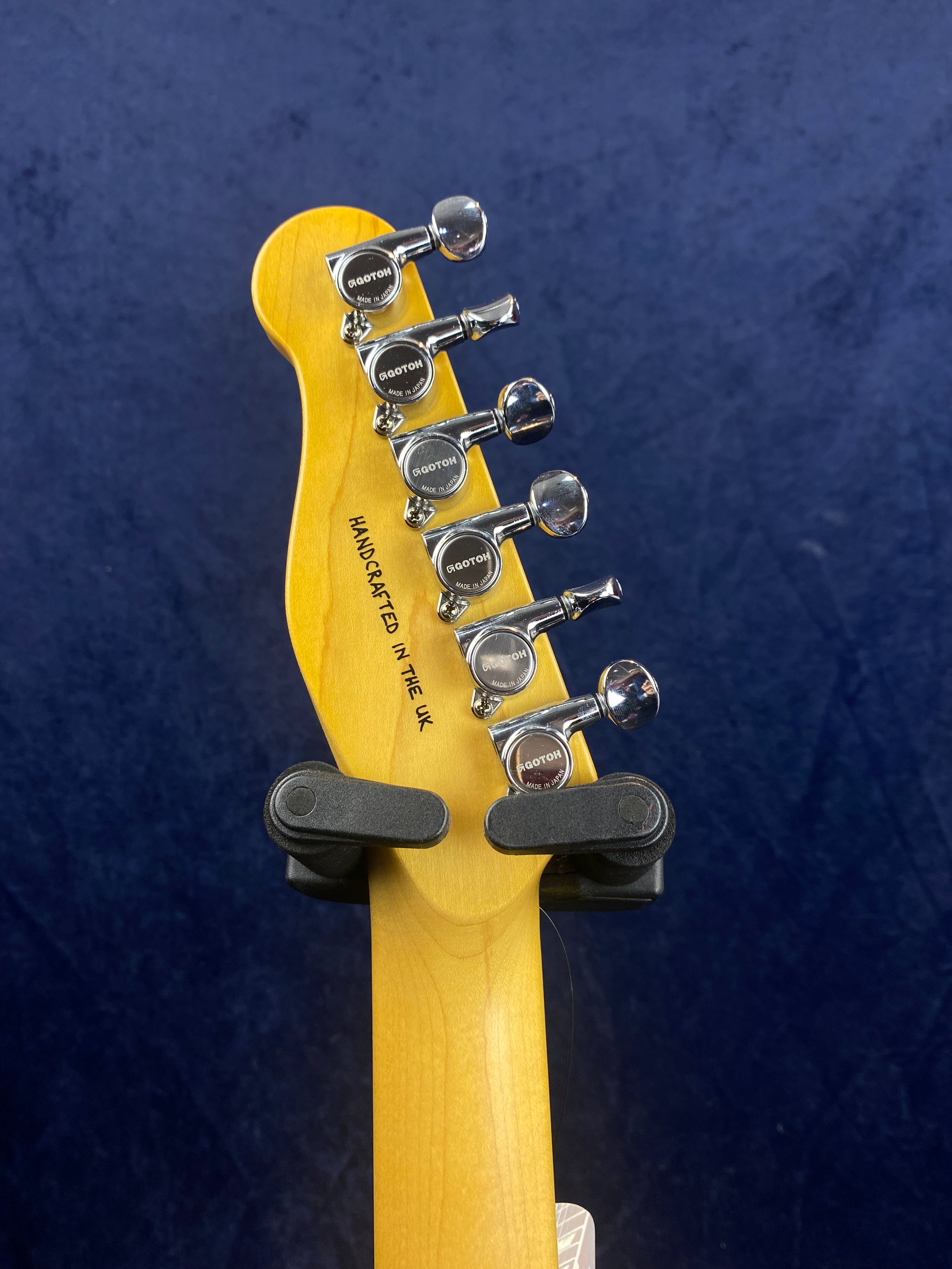 Six chrome Gotoh tuners on a light wood electric guitar headstock, “Handcrafted in the UK” text visible, held in a black guitar wall mount.