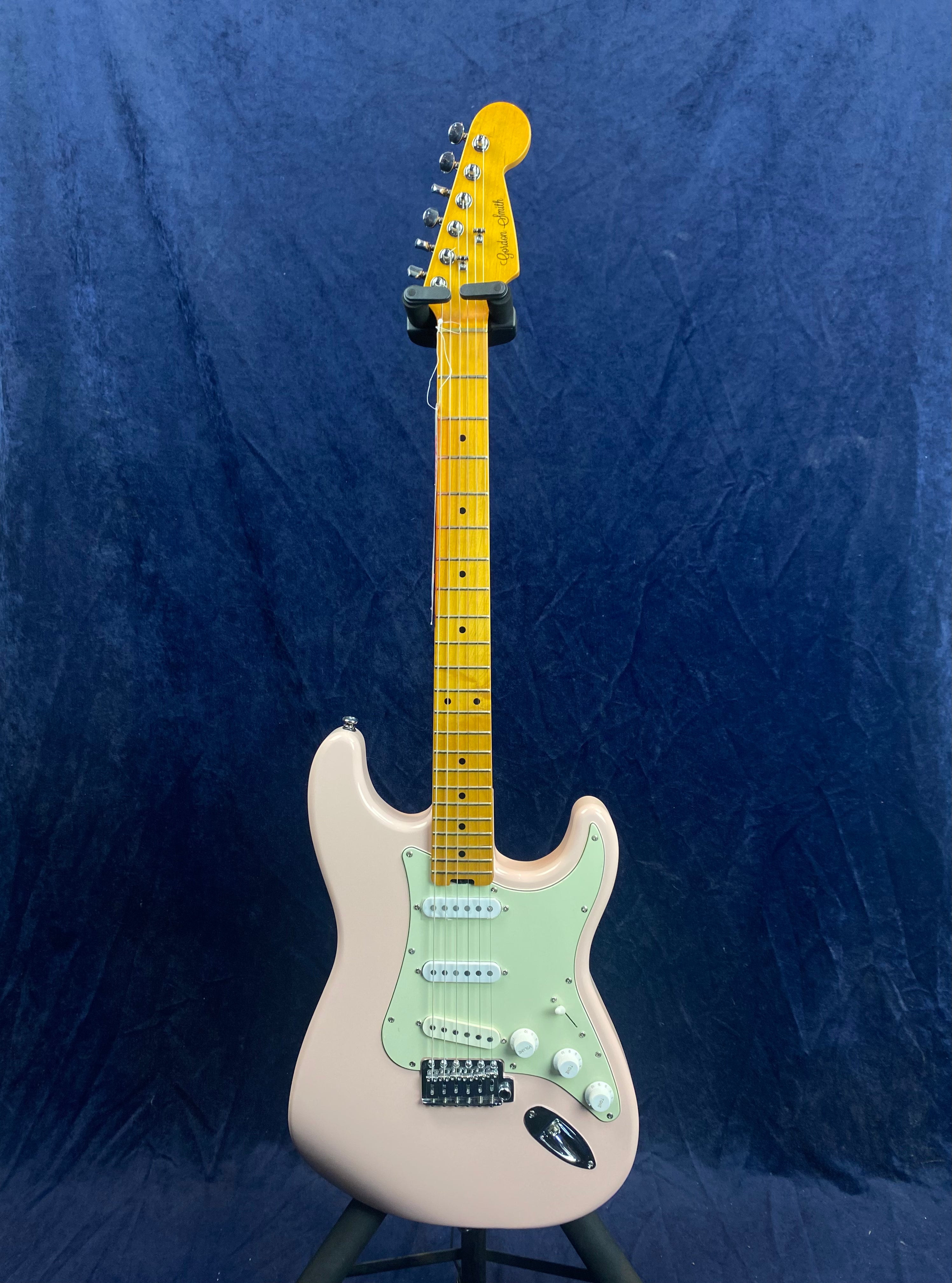 Light pink electric guitar with a maple neck, white pickguard, three single-coil pickups, and chrome hardware, shown upright against a dark blue background.