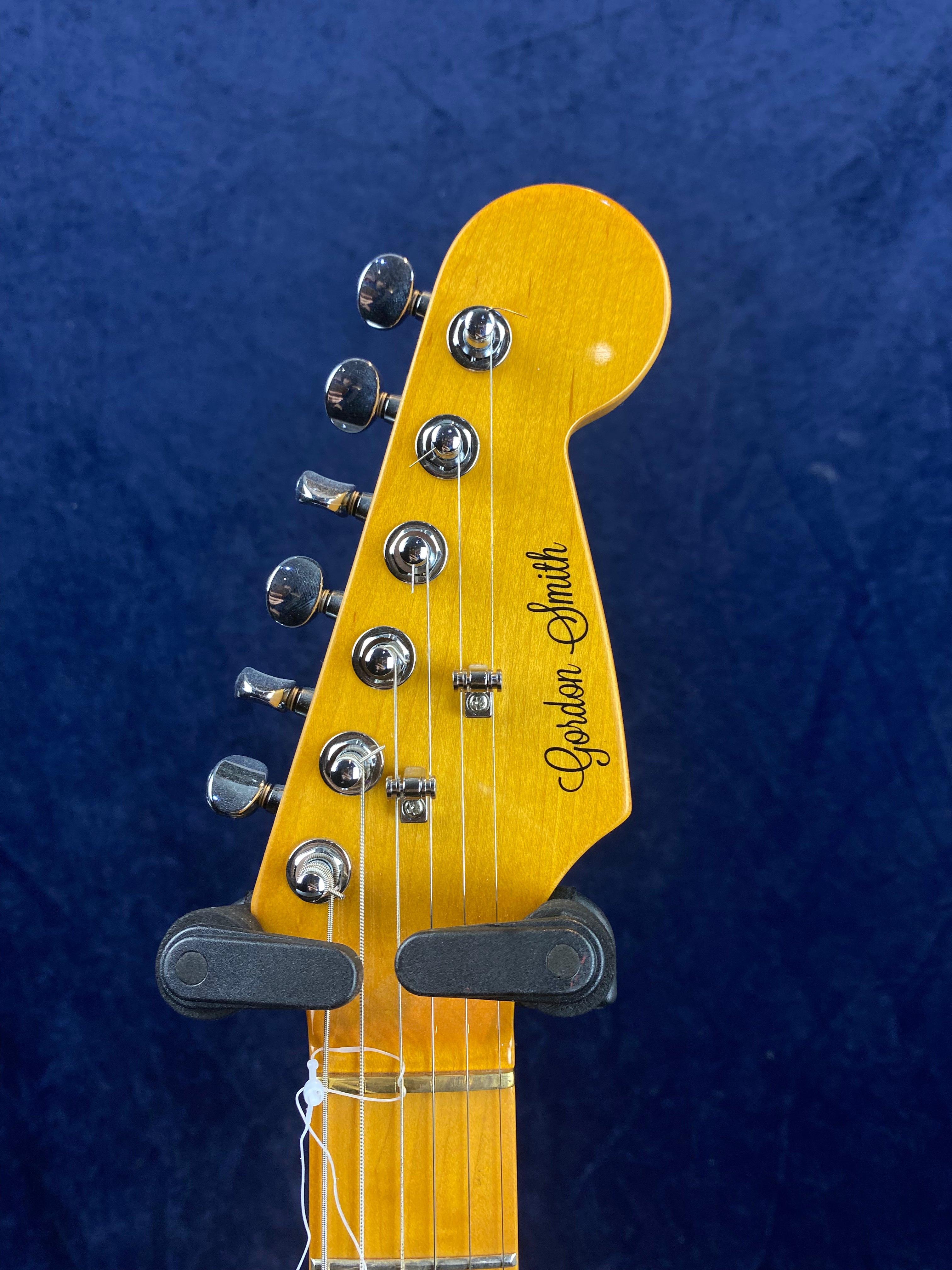 Yellow electric guitar headstock with six chrome tuning pegs, black "Gordon Smith" logo, and strings, held by a black wall mount against a dark blue background.