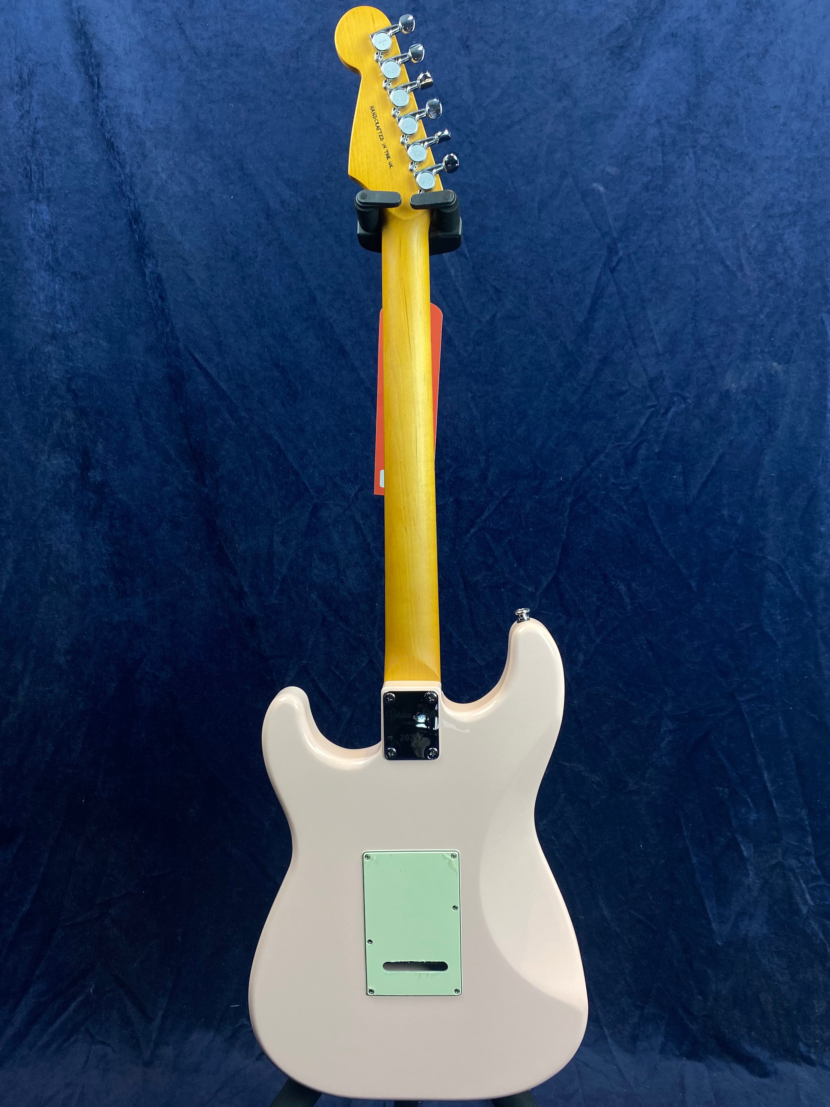 Electric guitar shown from the back with a light pink body, light green back plate, and natural wood neck against a dark blue background.