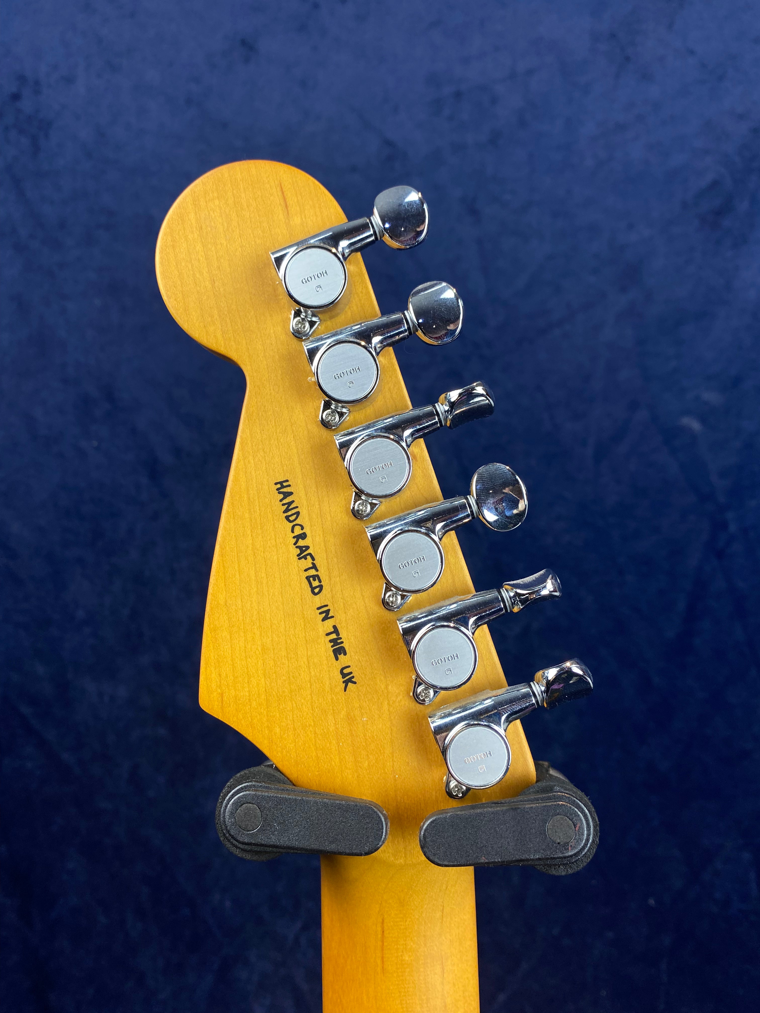 The back of an electric guitar headstock shows six chrome tuning pegs and the text "HANDCRAFTED IN THE UK" on natural wood.