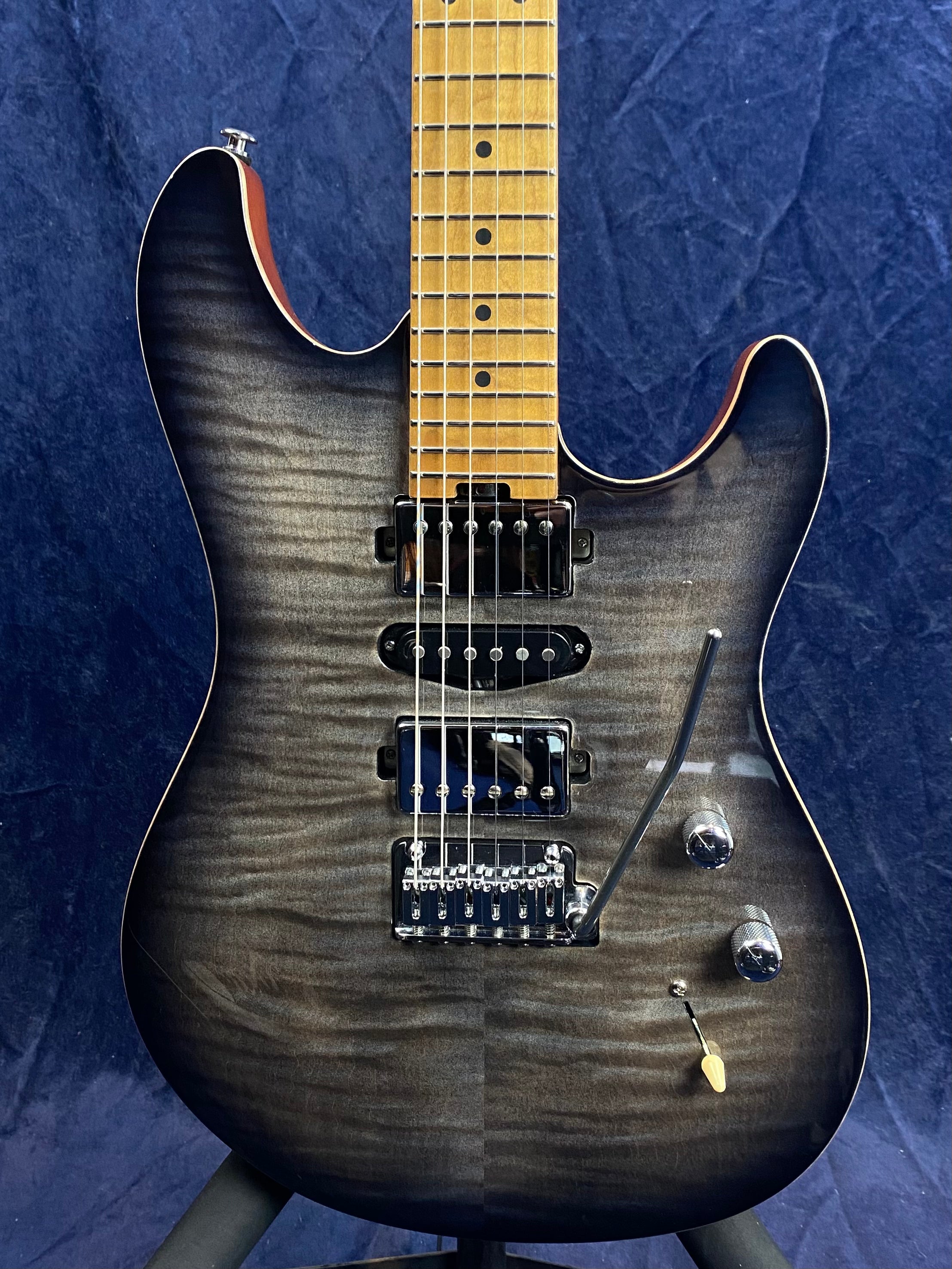 Electric guitar with a gray-black flame maple top, three pickups, two control knobs, a tremolo arm, and a light wooden neck with dot inlays.