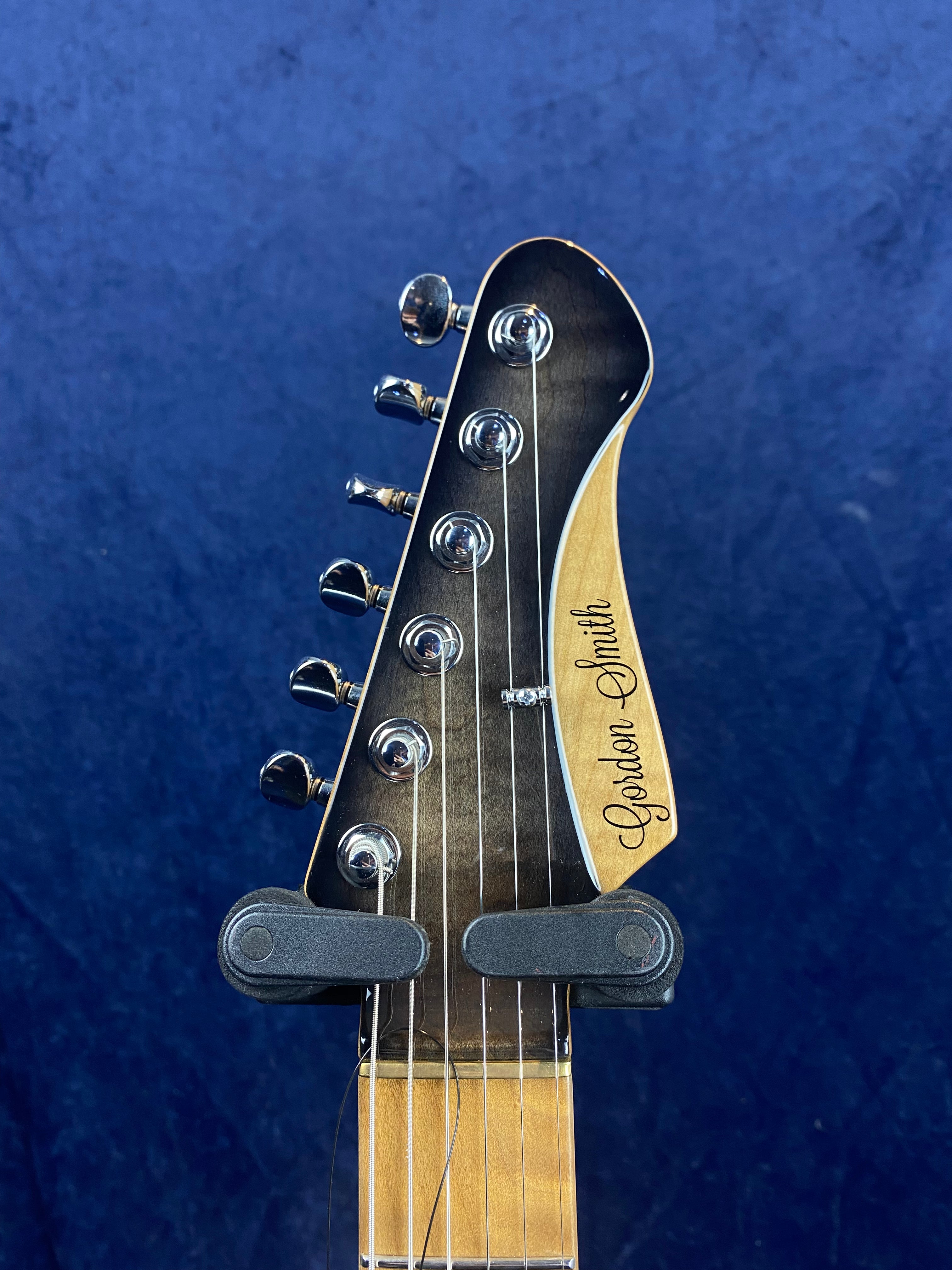 Six tuning pegs on a Gordon Smith electric guitar headstock, wood finish with black front, blue background.