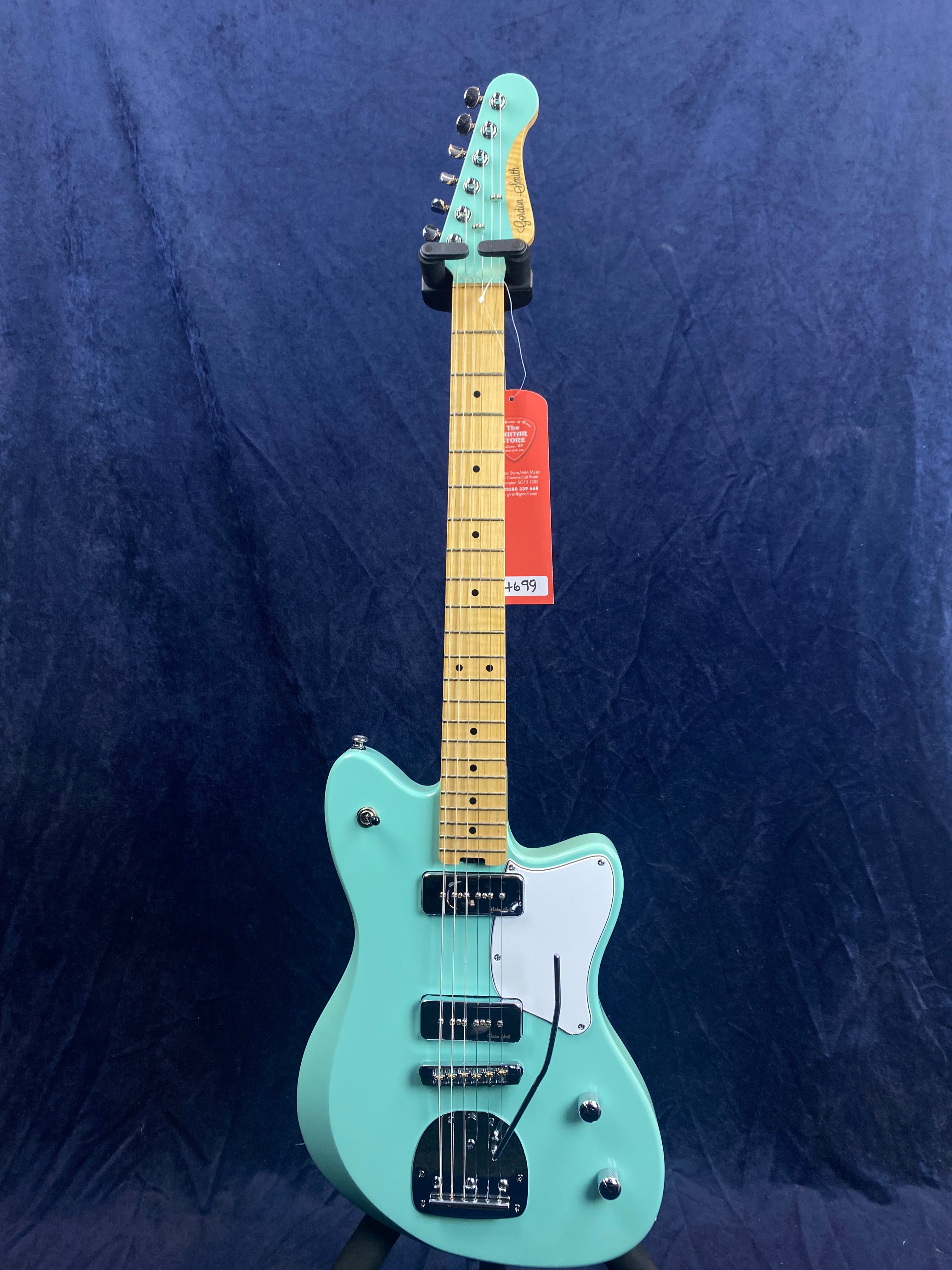 A turquoise electric guitar with a maple neck, two pickups, white pickguard, and chrome hardware is displayed upright against a dark blue background.