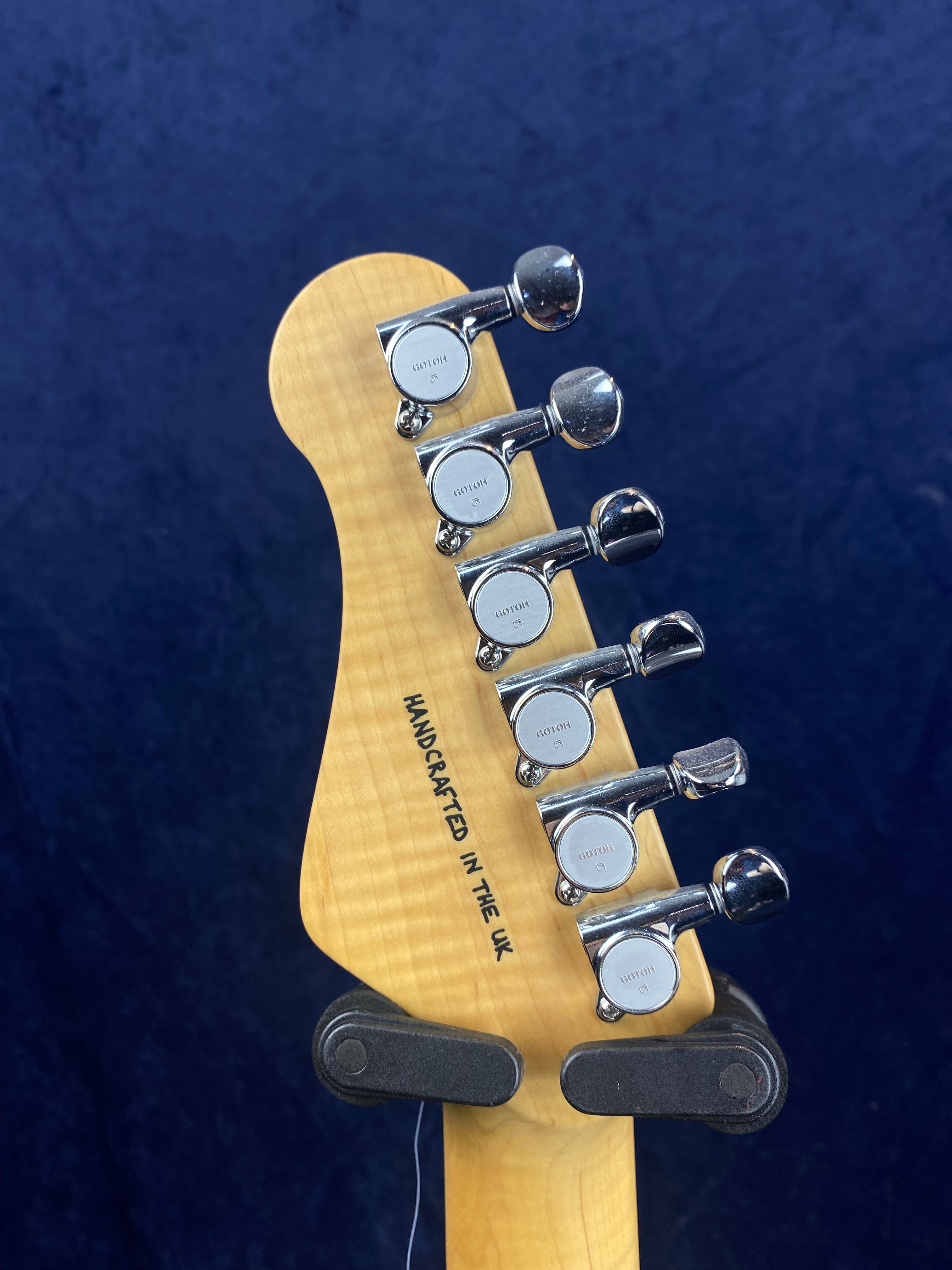 Electric guitar headstock with six chrome tuning pegs and "HANDCRAFTED IN THE UK" text on light wood, against a dark blue background.