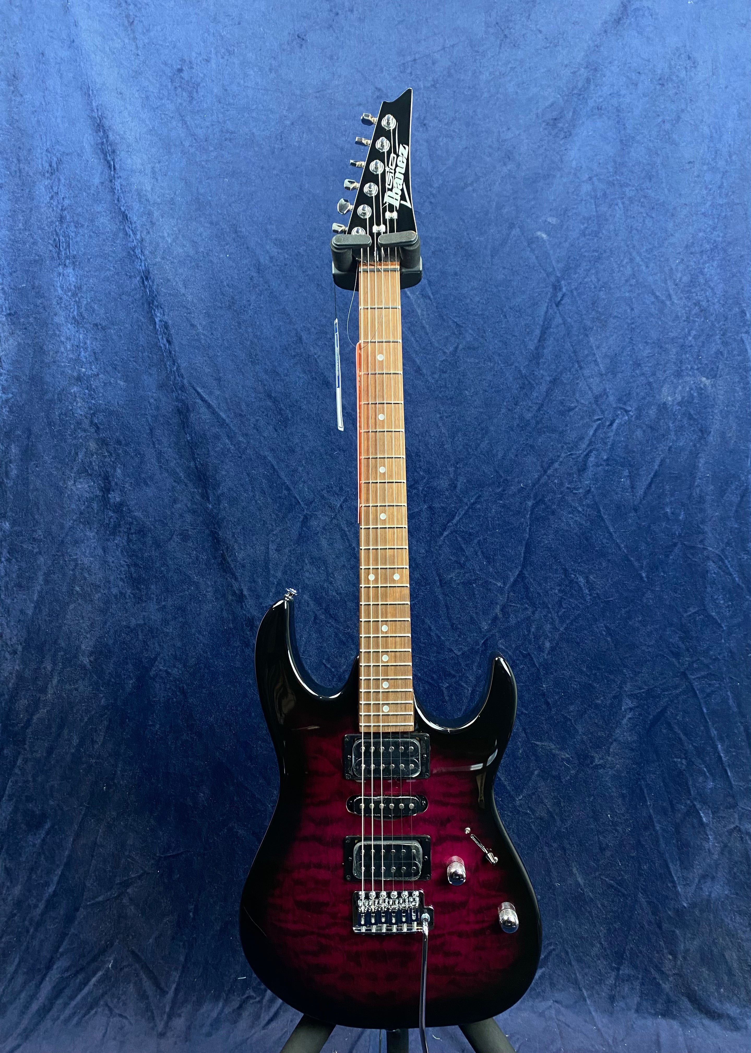Electric guitar with a dark red and black contoured body, three pickups, two control knobs, and a light brown fretboard against a blue background.