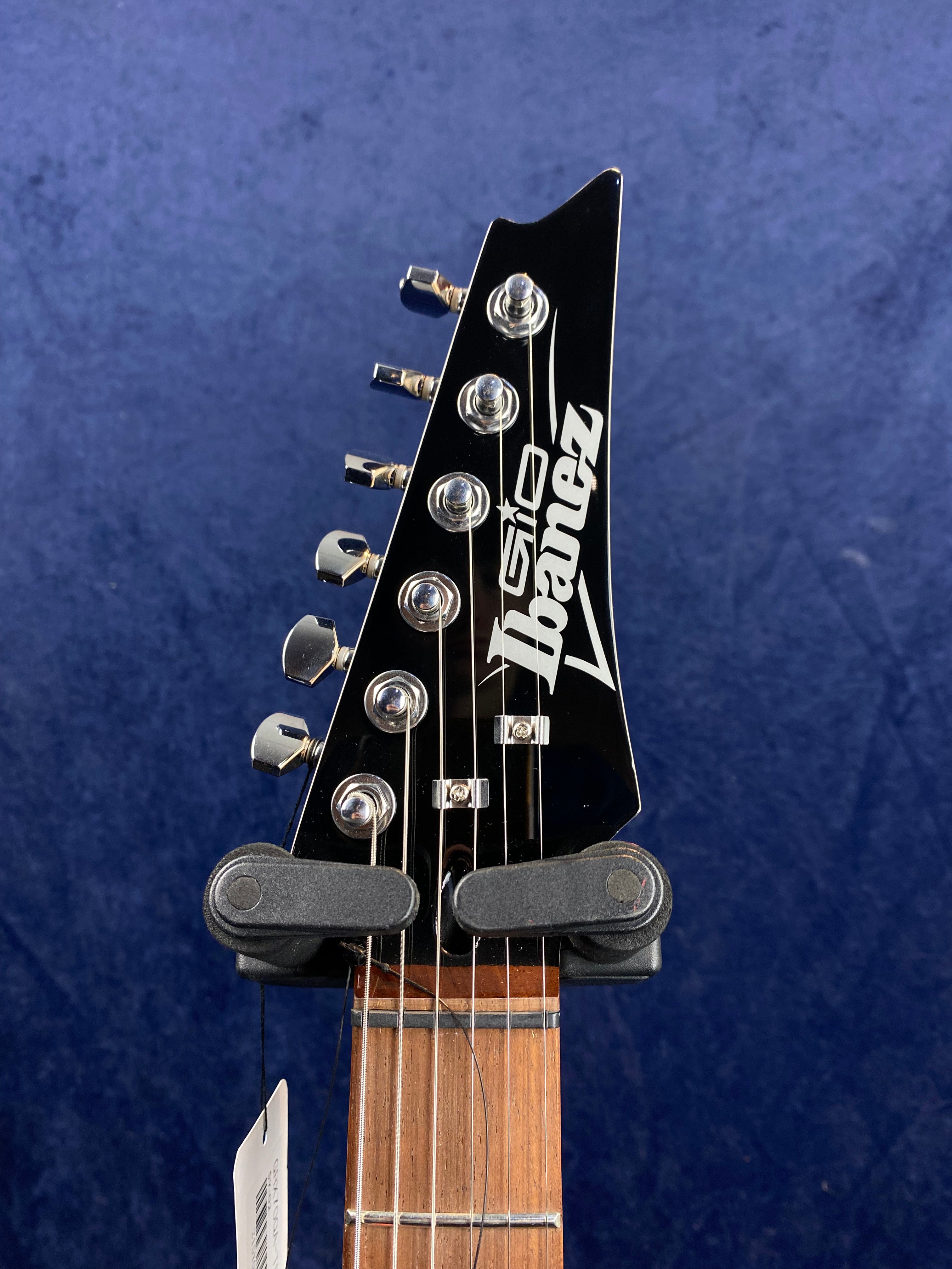 Ibanez electric guitar headstock with six chrome tuning pegs, black finish, and bold white Ibanez logo, against a dark blue background.