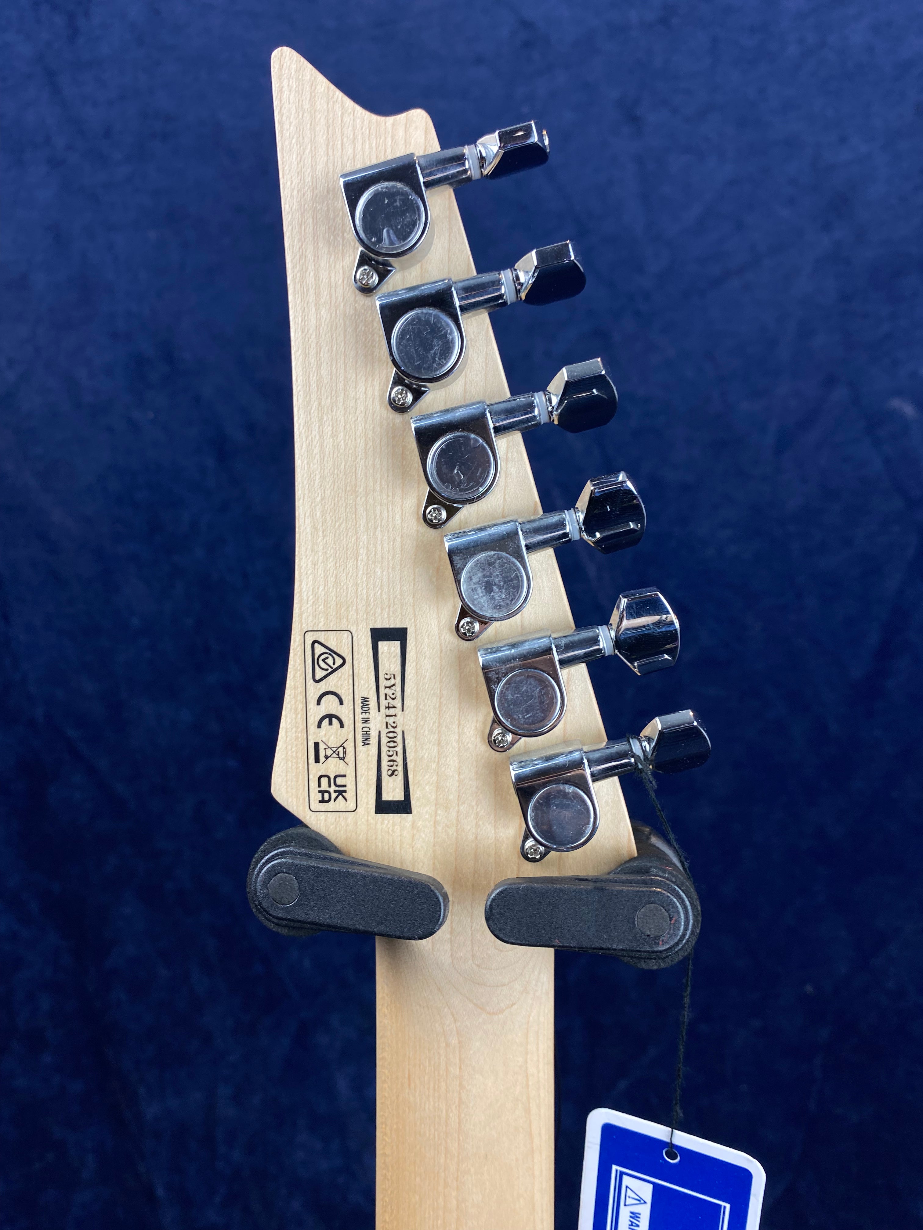 Six chrome tuning pegs on a natural wood electric guitar headstock, visible manufacturer markings, and a blue warning tag hanging from the neck.