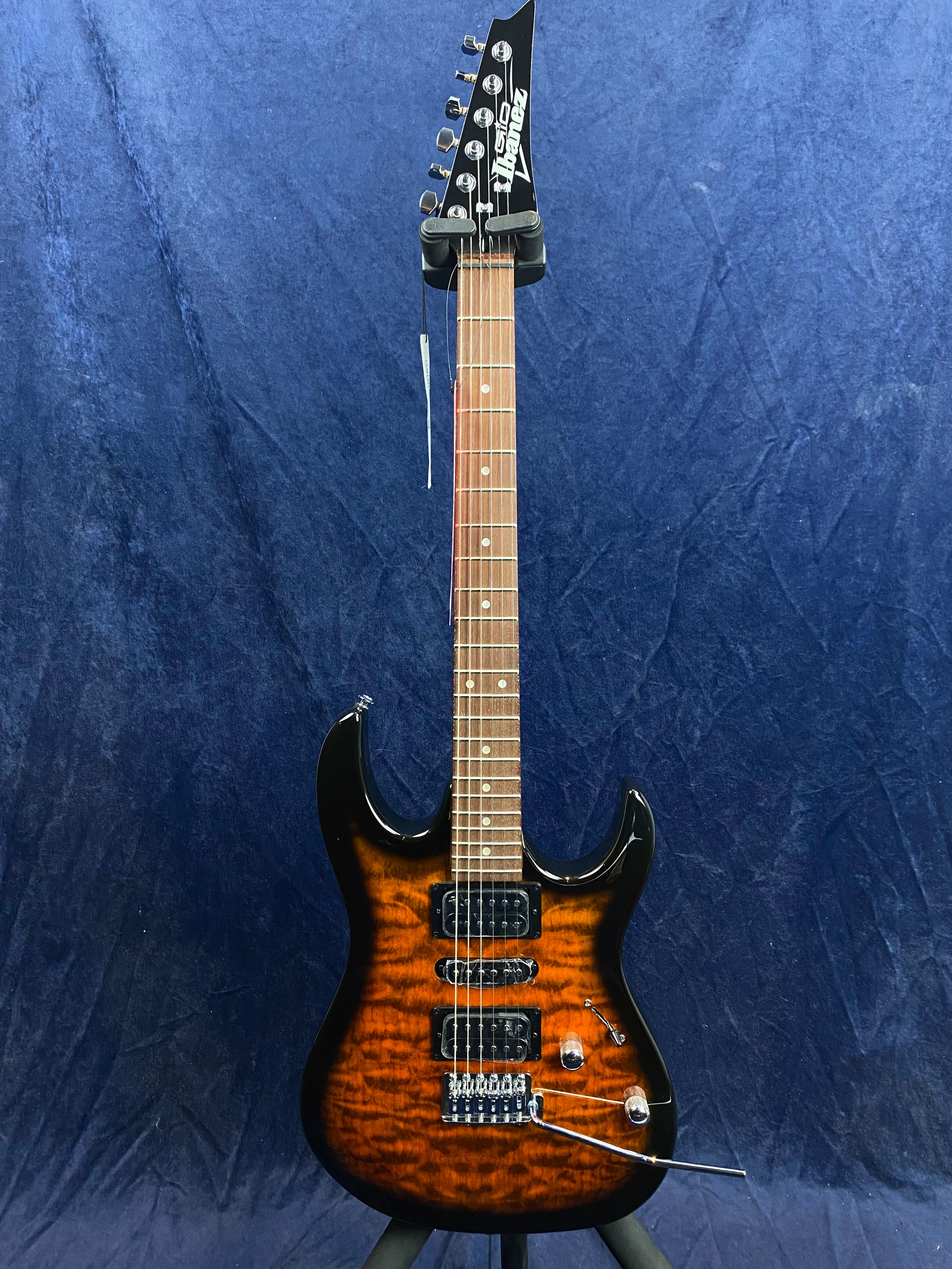 Ibanez electric guitar with a dark sunburst finish, two humbucker pickups, one single-coil pickup, and a tremolo bar, against a blue background.