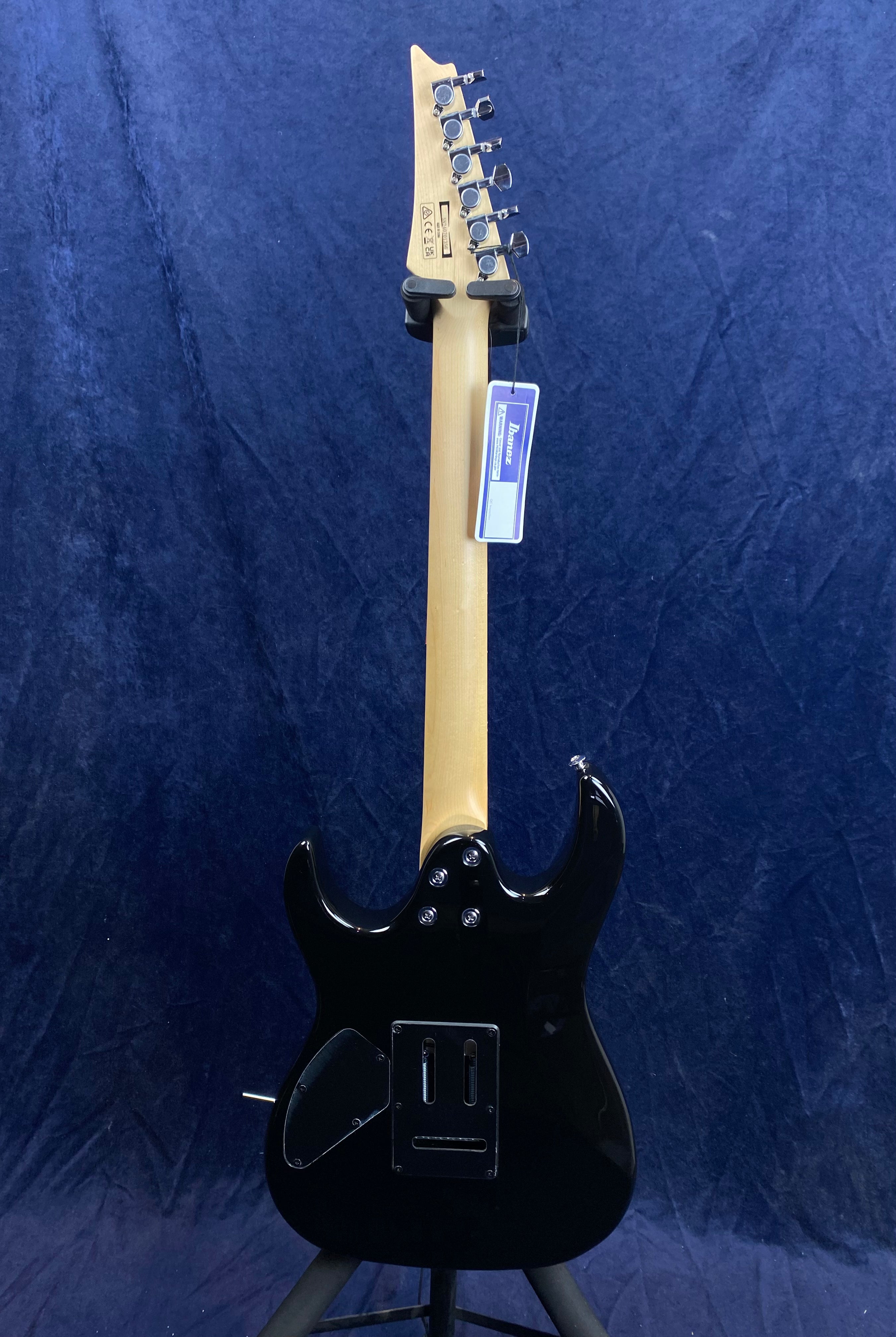 Black electric guitar shown from the back, light wood neck, six tuning pegs, Ibanez tag hanging from the headstock, two rear cover plates visible.