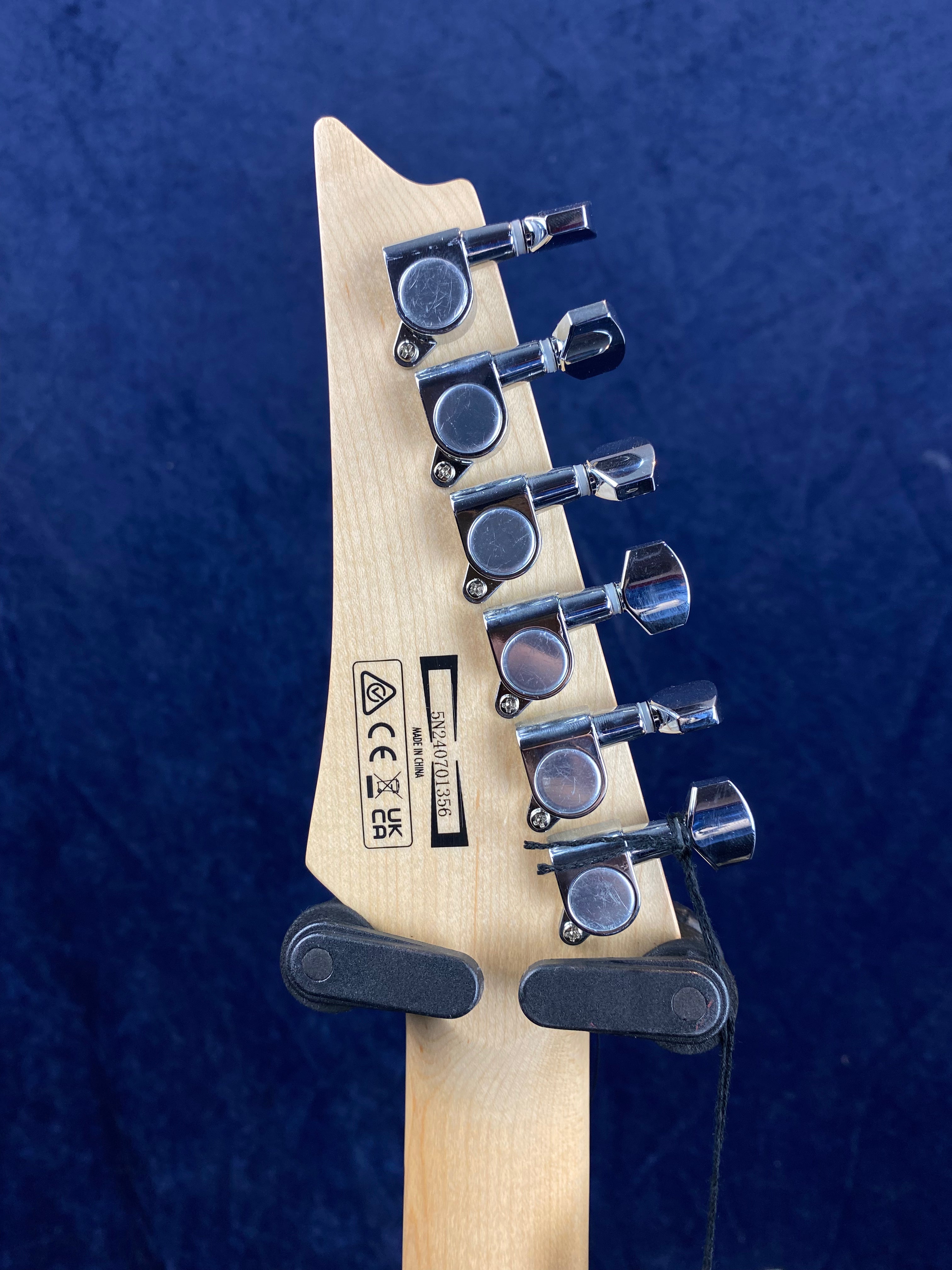 Six chrome tuning pegs on a light wood electric guitar headstock, with manufacturer labels and serial number visible above a black wall mount.